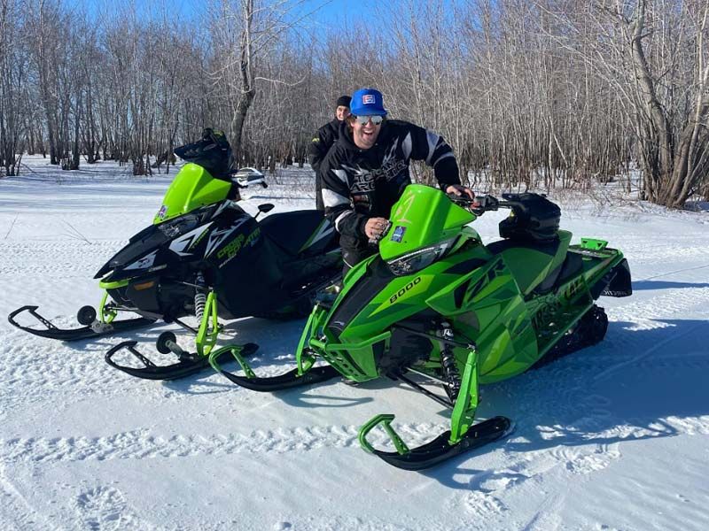 a man in a blue hat stands next to two green snowmobiles