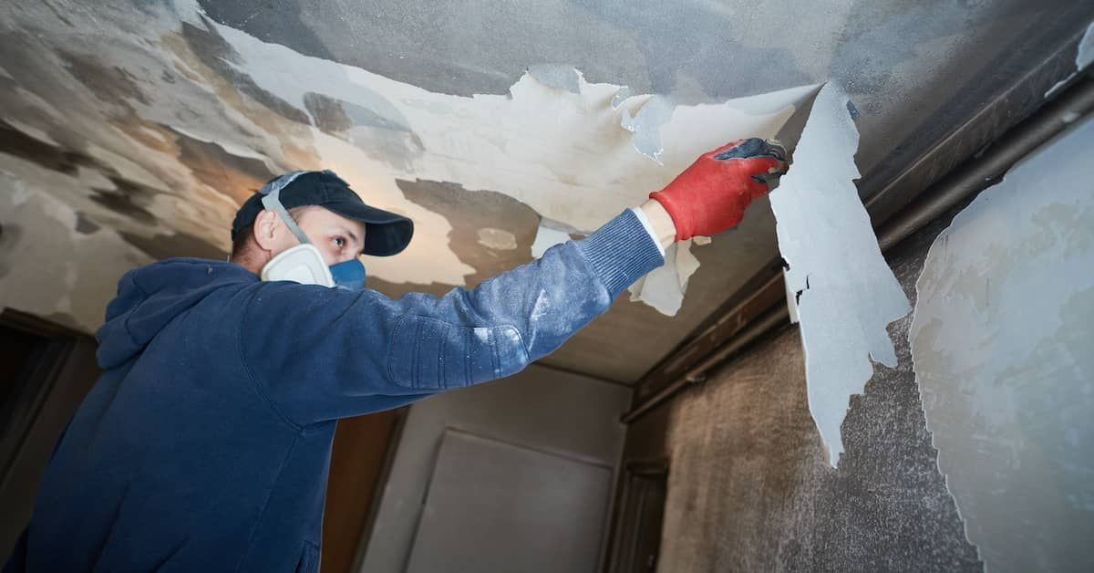 Man removing peeling plaster from a ceiling wearing a respirator and gloves in a room.