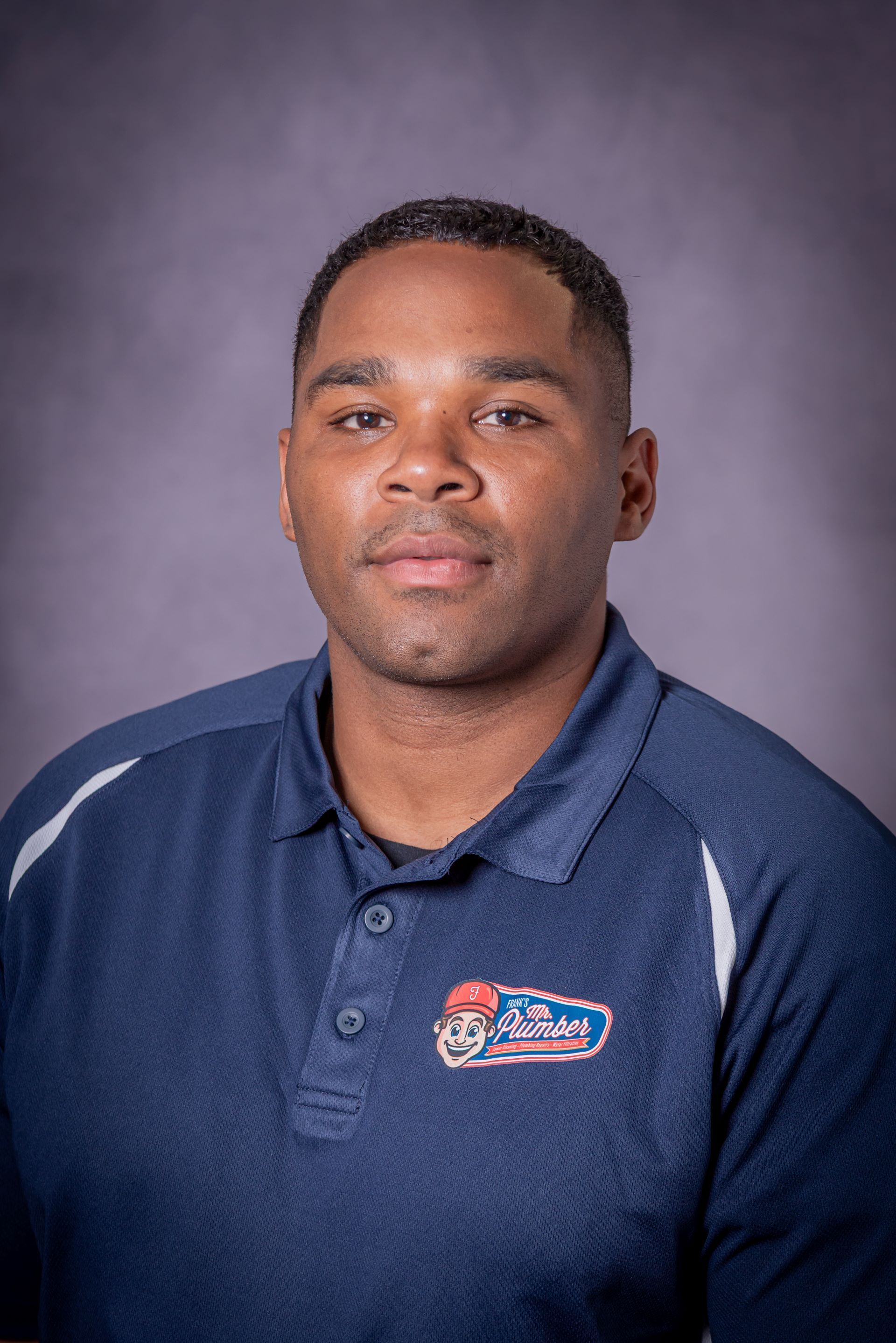 A man in a blue polo shirt is standing in front of a purple background.