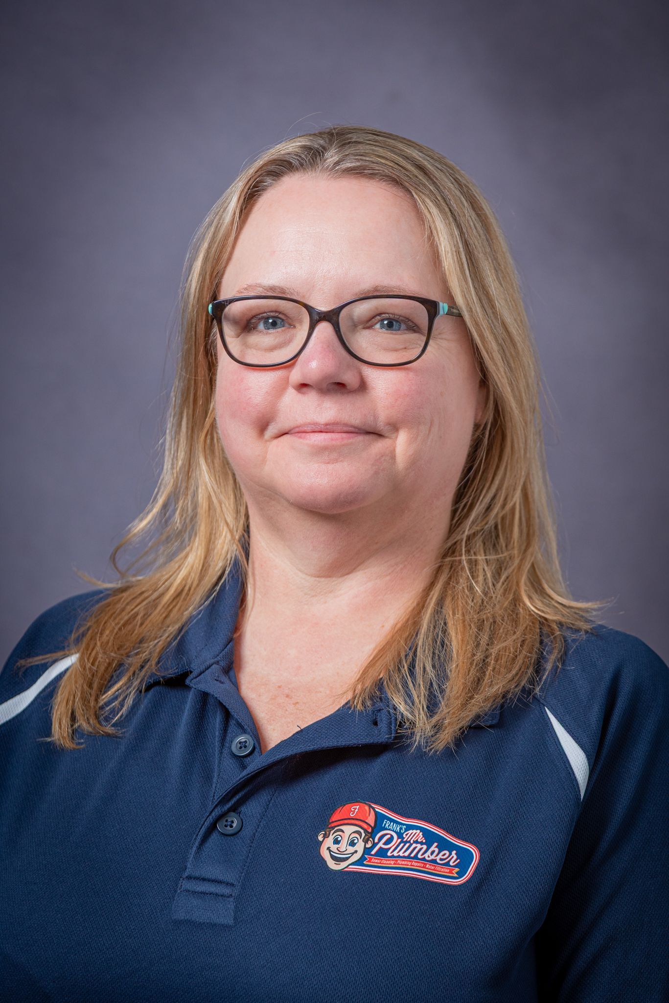 Woman with blonde hair and glasses wearing a navy blue polo shirt with a logo, smiling, on a gray background.