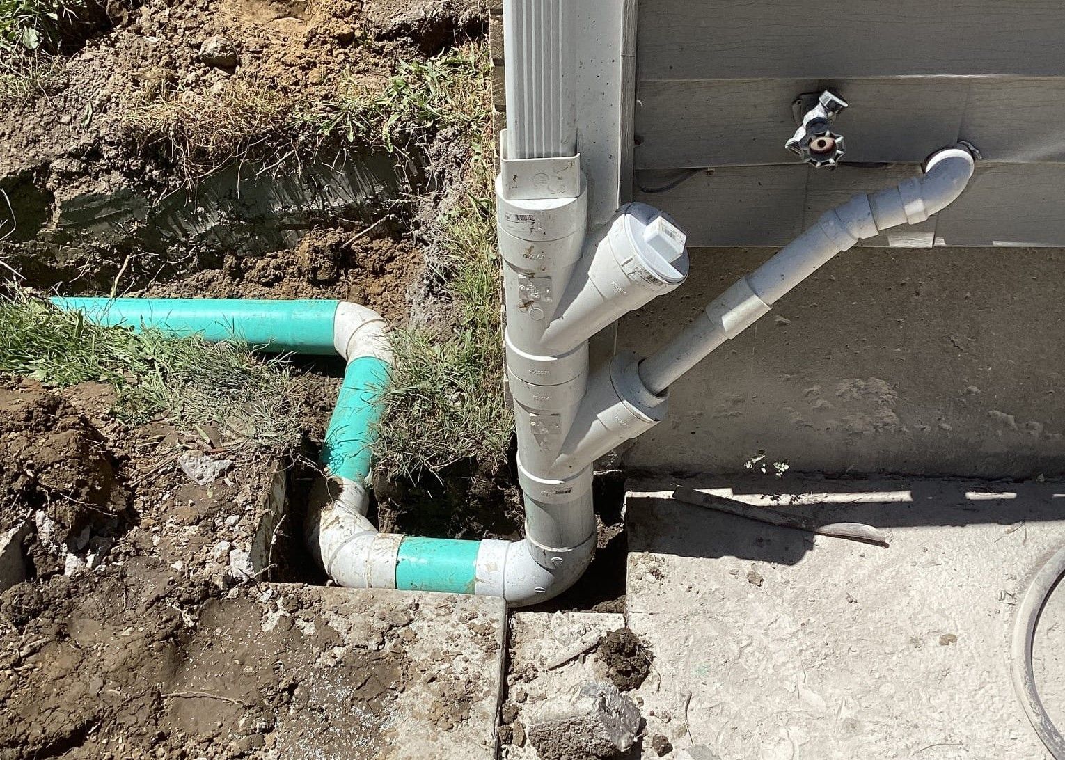 Drainage pipes directing water away from a house foundation. Turquoise and gray pipes in the dirt.