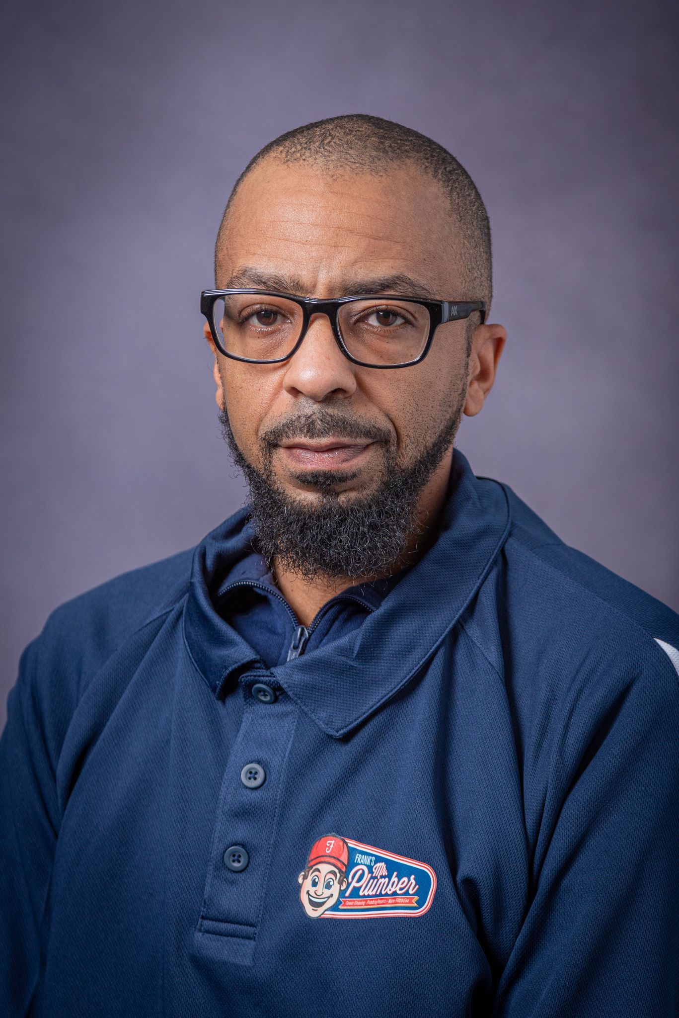 Man with glasses and a beard wearing a blue shirt with a logo, against a purple backdrop.