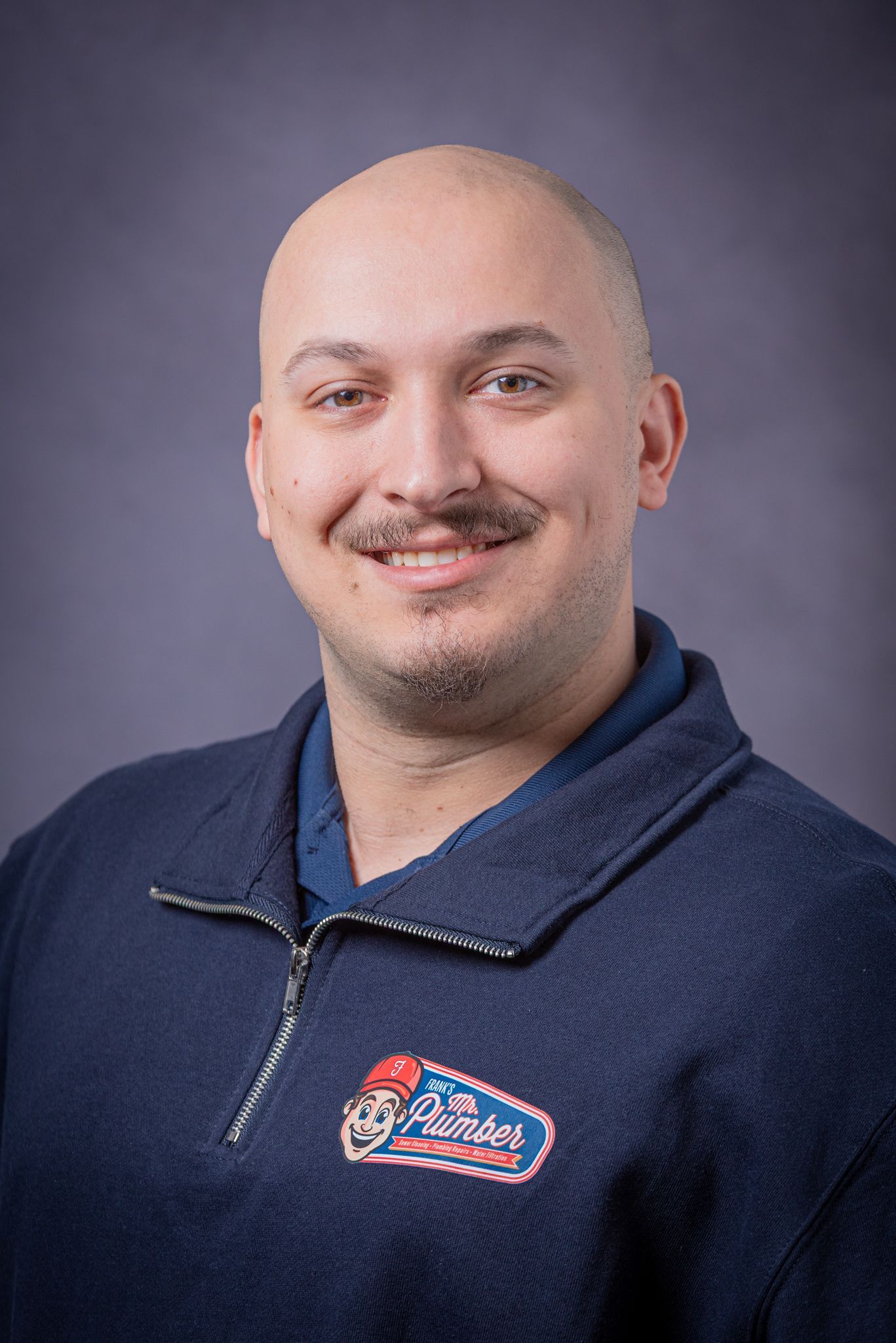 Bald man with mustache smiles, wearing a navy blue sweater with logo patch.