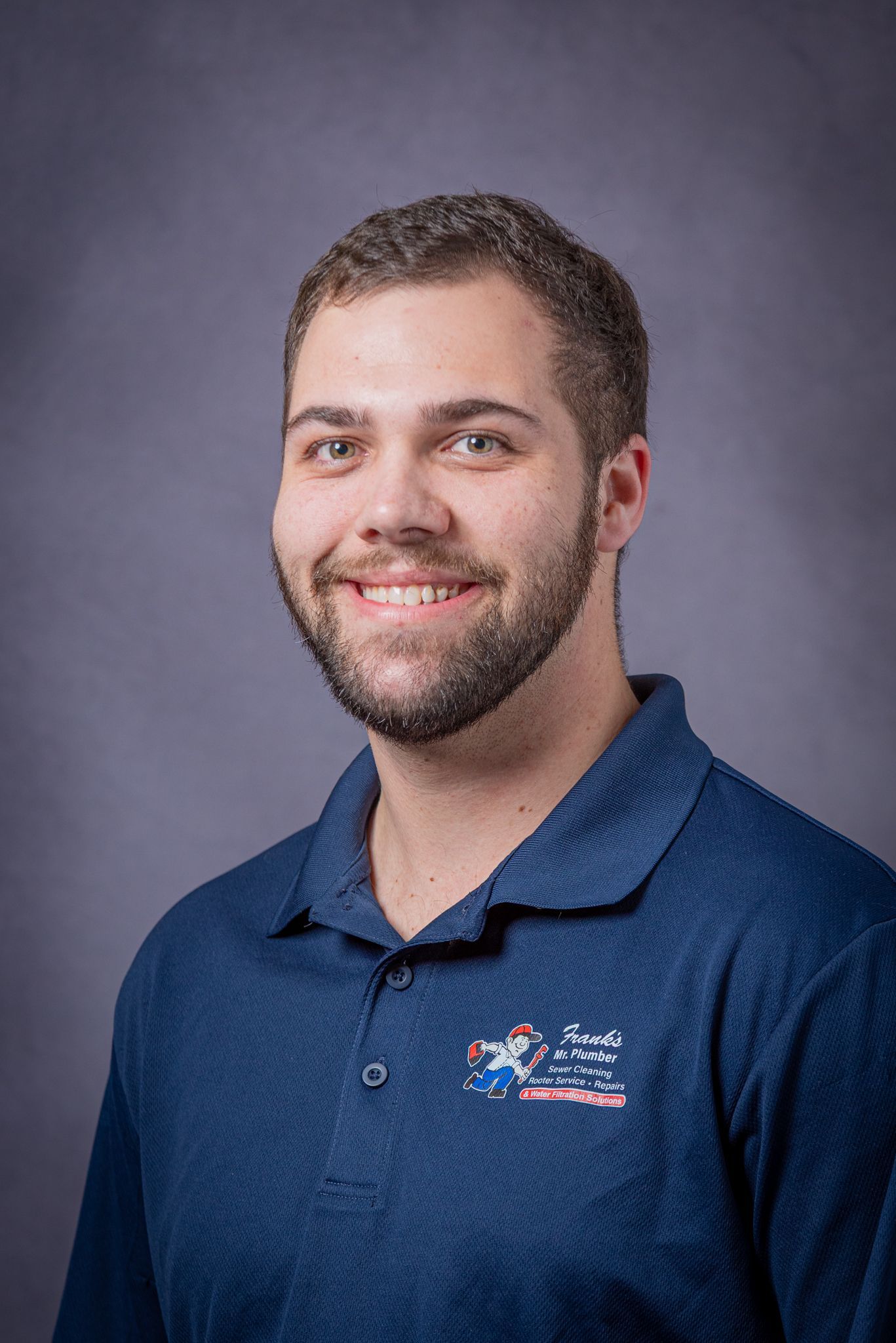 Man in blue polo shirt with logo, smiling against a purple backdrop.