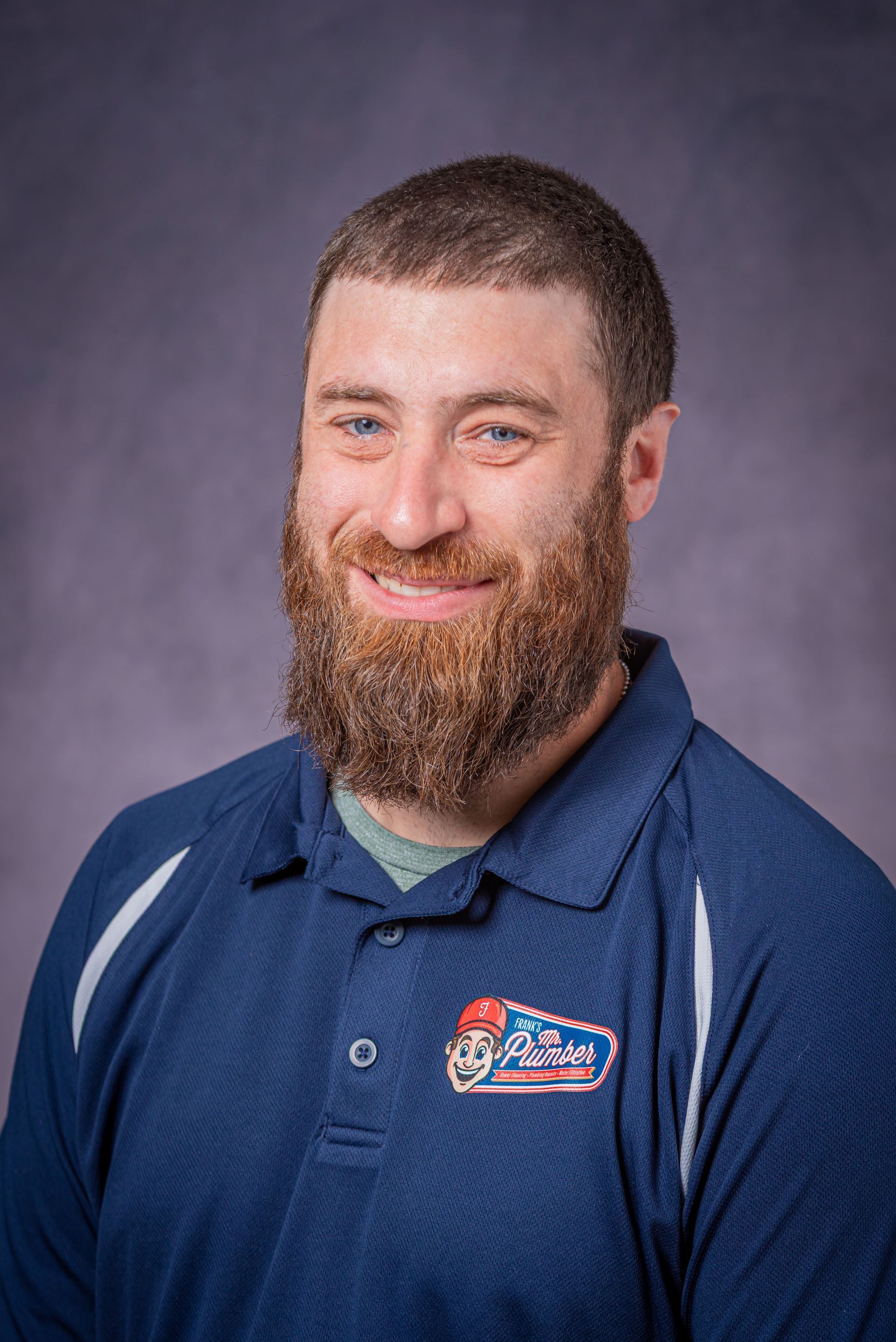 A man with a beard is wearing a blue polo shirt and smiling.