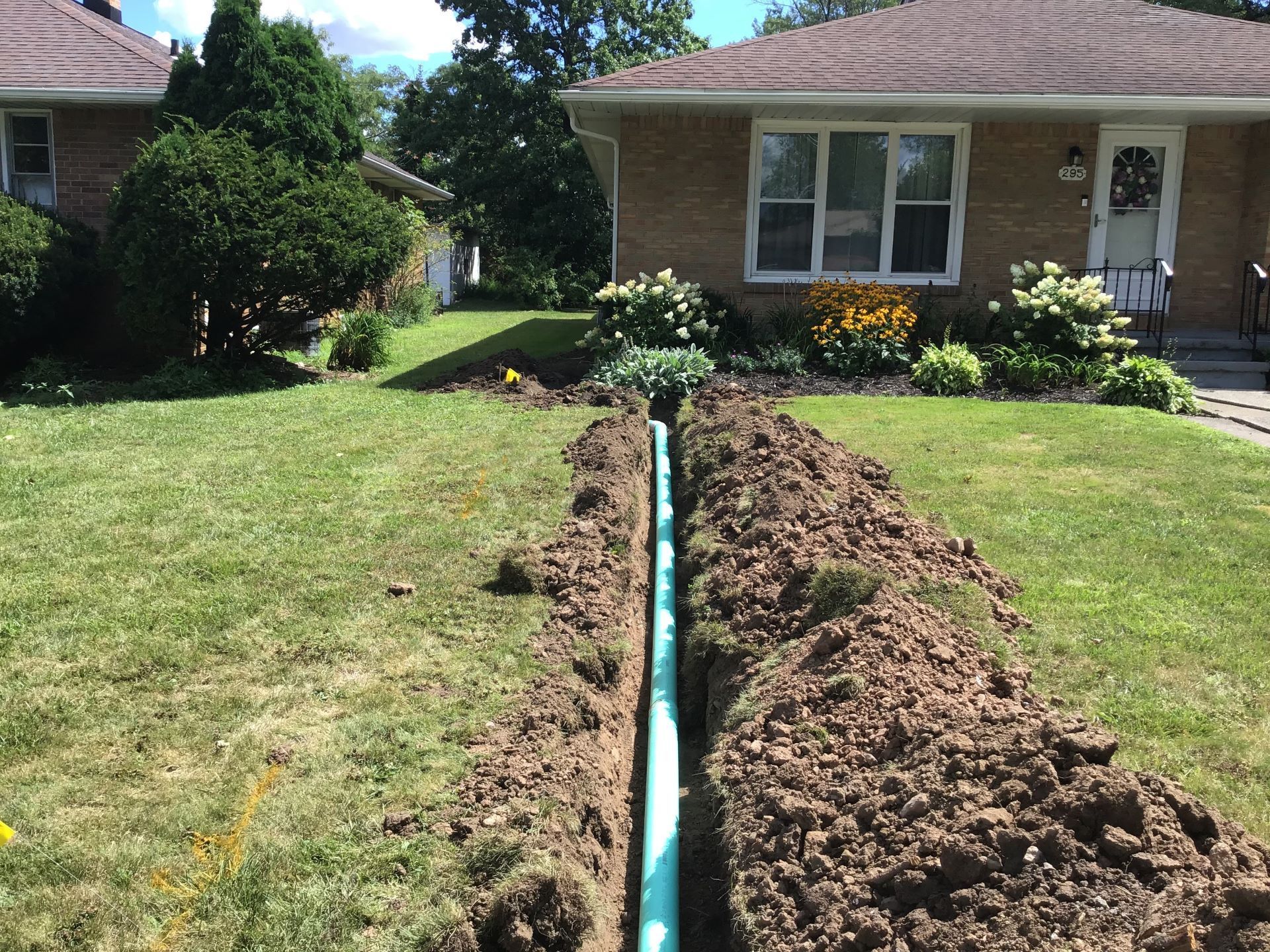 A trench in a front yard with exposed blue pipe; house and landscaping in the background.