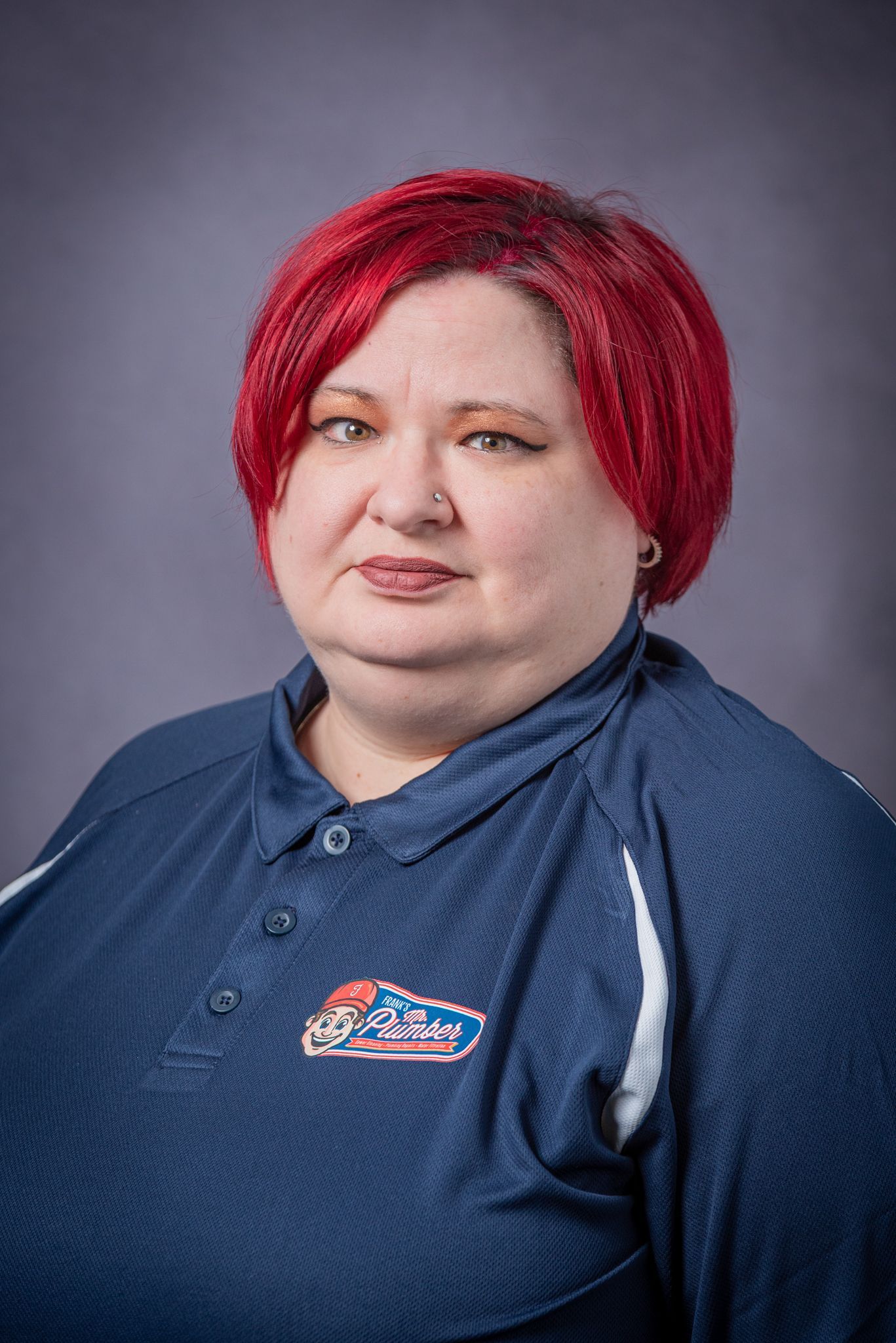 Woman with red hair, wearing a navy polo shirt with logo, stands in front of a gray backdrop.