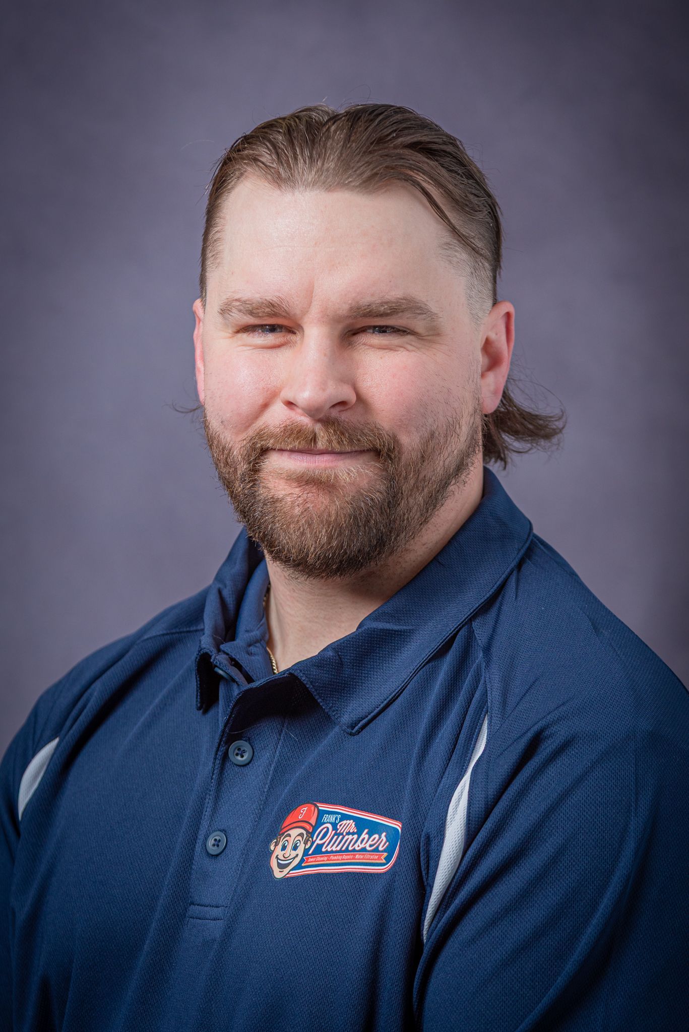 Man with a beard and ponytail wearing a blue polo shirt, smiling slightly.