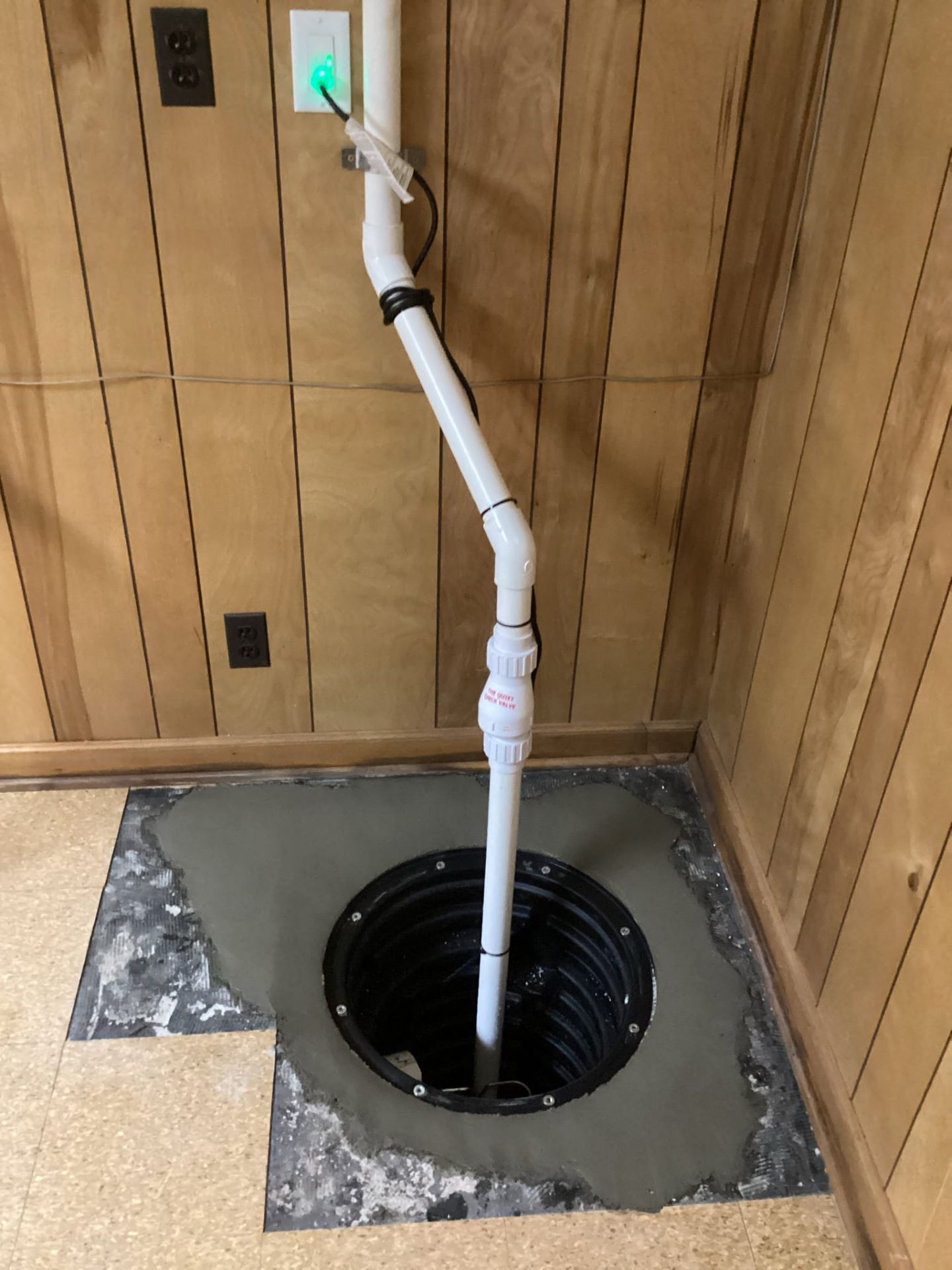 Sump pump in basement floor with white PVC pipe, surrounded by concrete and wood paneling.