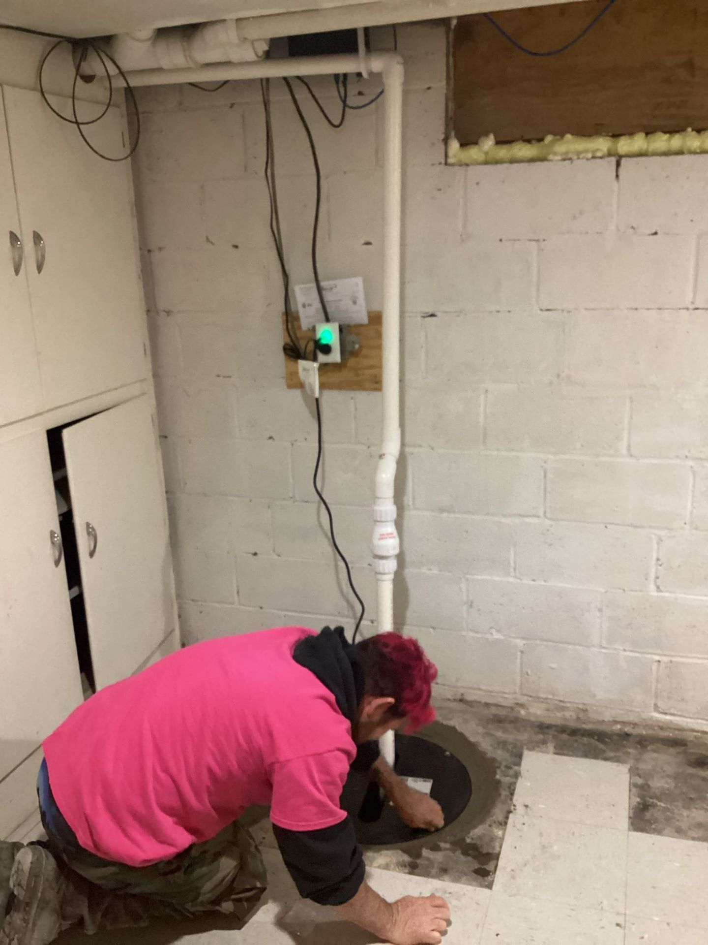 Person in pink shirt works on sump pump in a basement with white block walls and a storage cabinet.