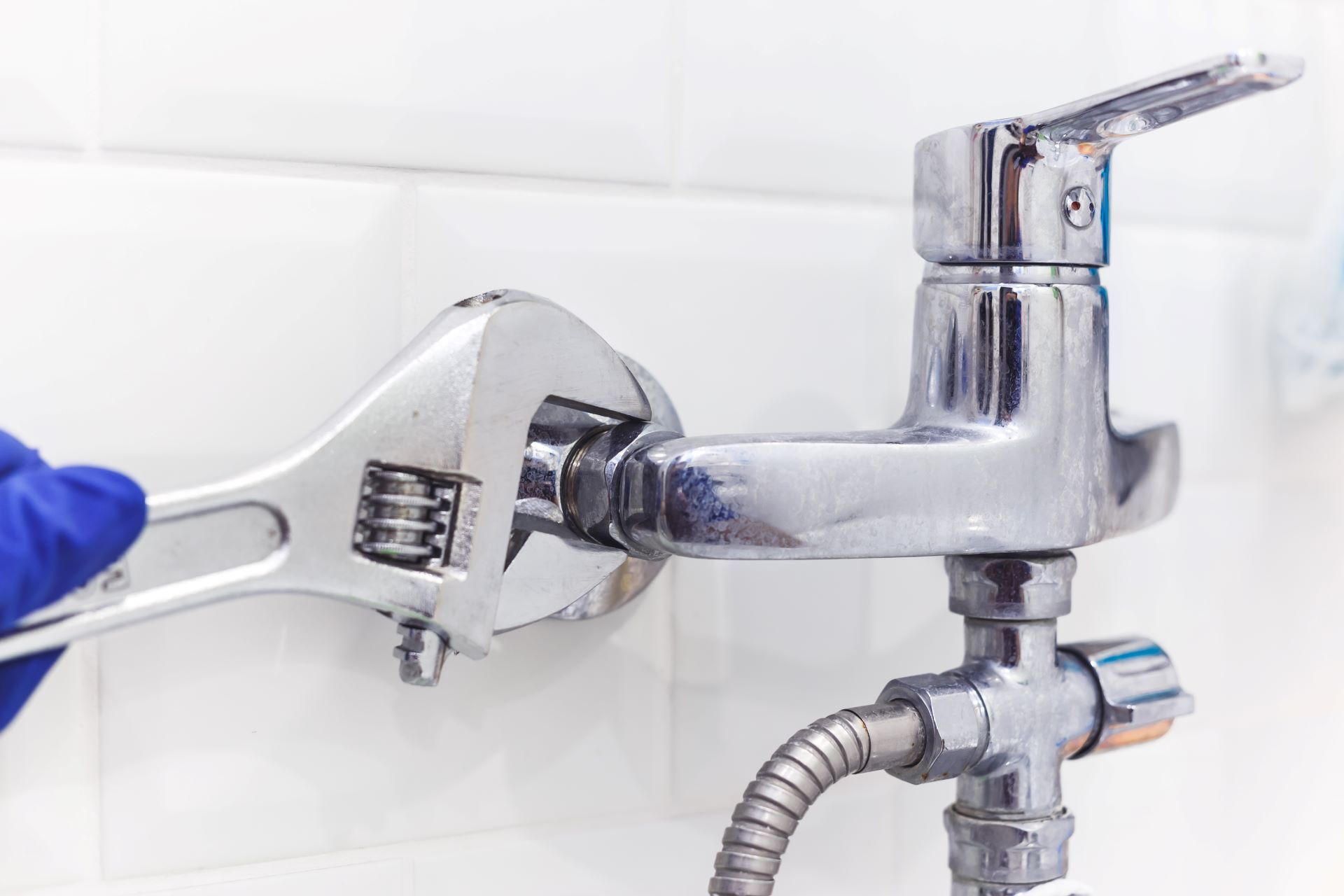 A person with a gloved hand uses a wrench to tighten a faucet against a tiled wall.