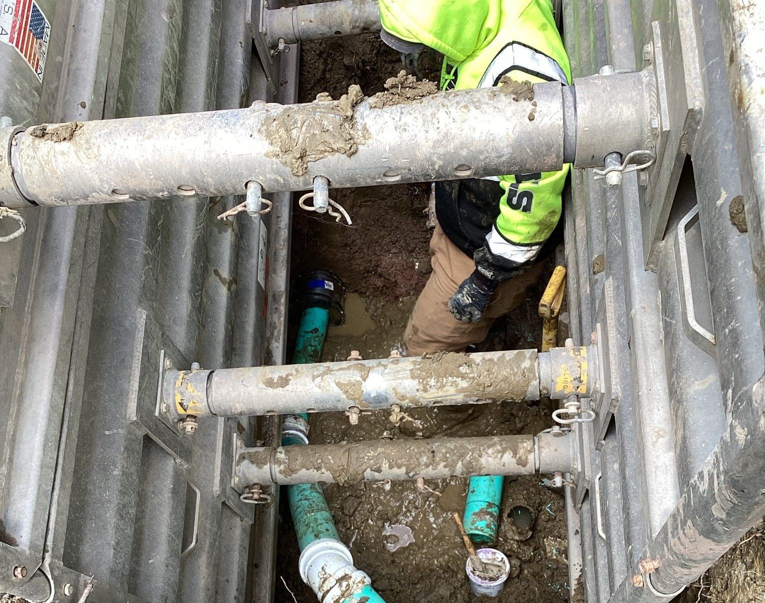 Worker in trench, wearing safety vest, connecting pipes. Trench box provides protection.
