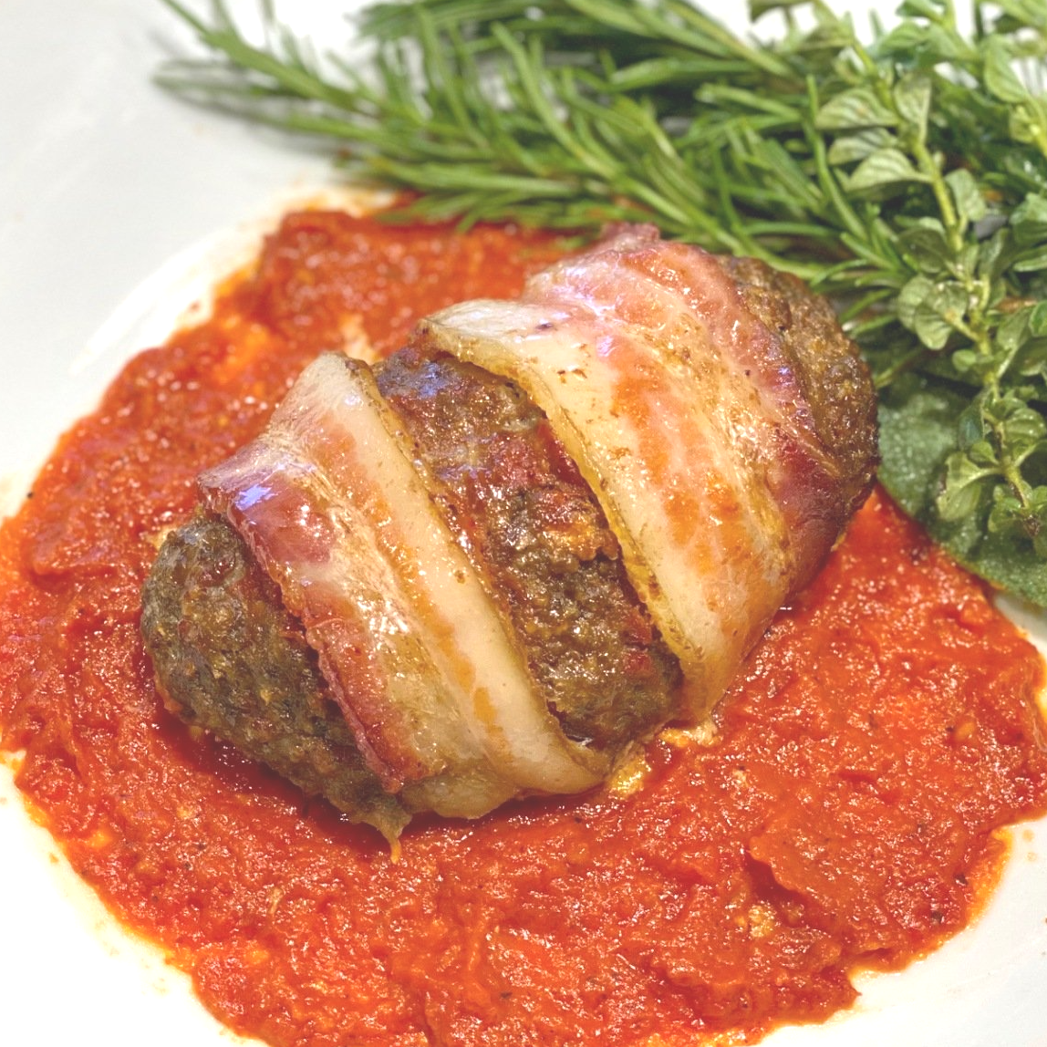 A meatball wrapped in bacon is on a plate with tomato sauce.
