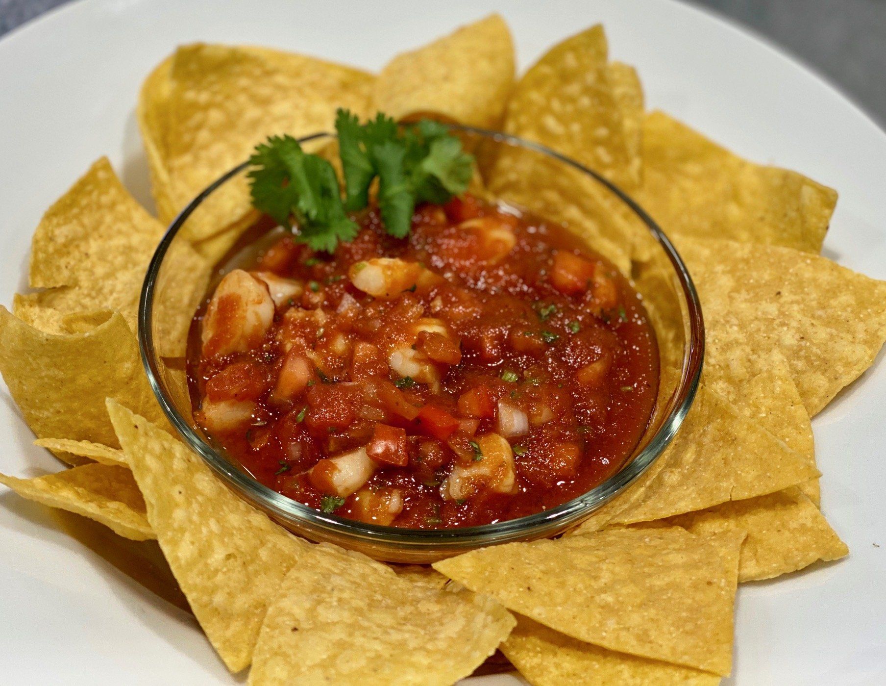 A bowl of salsa is surrounded by tortilla chips on a white plate.