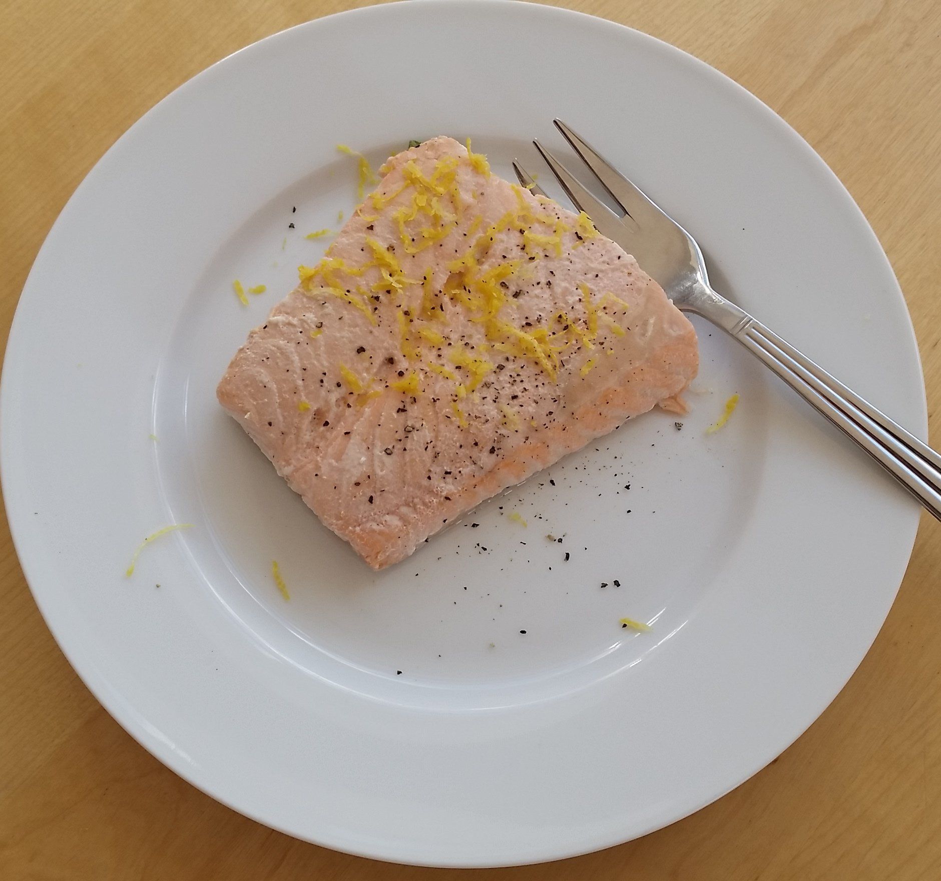 Cooked salmon fillet on a white plate, seasoned with black pepper and lemon zest. A fork rests beside it.