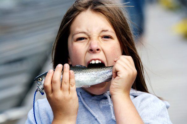 Girl grimaces while attempting to bite a fish. Outdoors, light skin, gray hoodie.