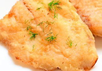 Fried fish fillet, golden-brown and garnished with fresh green dill, on a white plate.