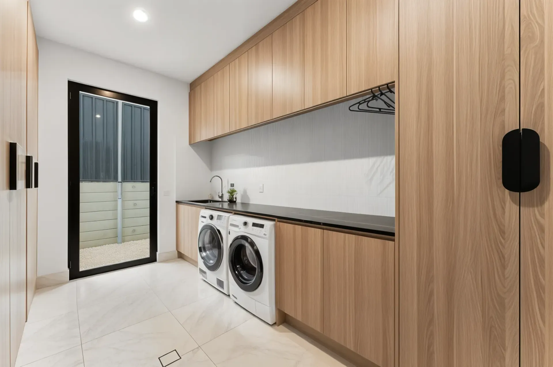Laundry room with light wood cabinets, white appliances, black countertop, and an exterior door — Caprice Cabinets in Coolum Beach, QLD