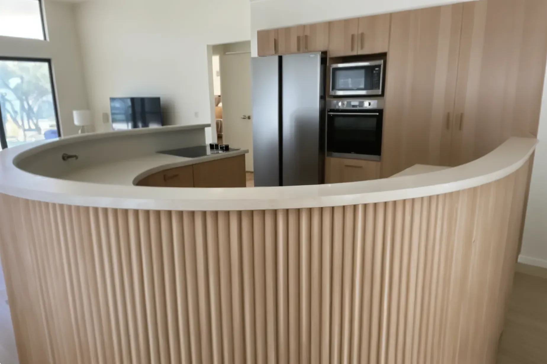Curved kitchen island with vertical wood slats. Built-in oven, microwave, and refrigerator in a light wood kitchen. — Caprice Cabinets in Maroochydore, QLD