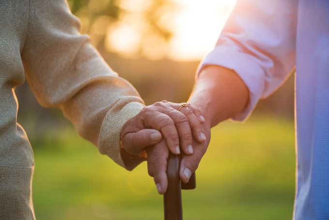 Elderly couple holding hands, one using a cane, outdoors in the sunlight.