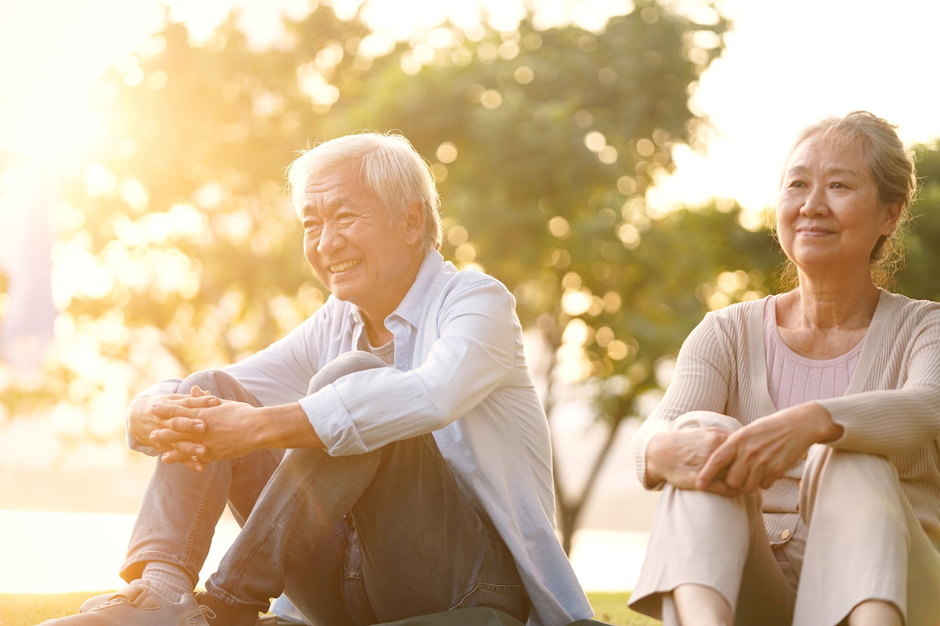Smiling elderly Asian couple sitting outdoors, bathed in sunlight.