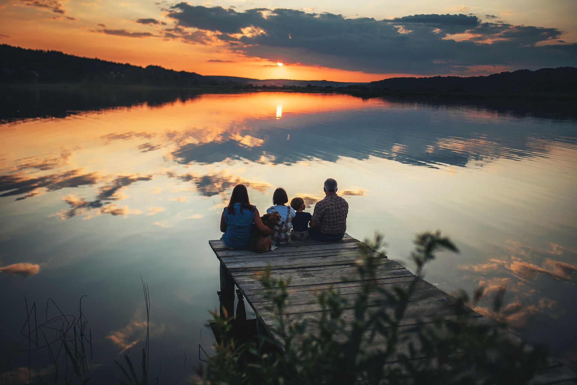 Family sitting on a dock, watching sunset over a calm lake. Golden sky reflects in the water.
