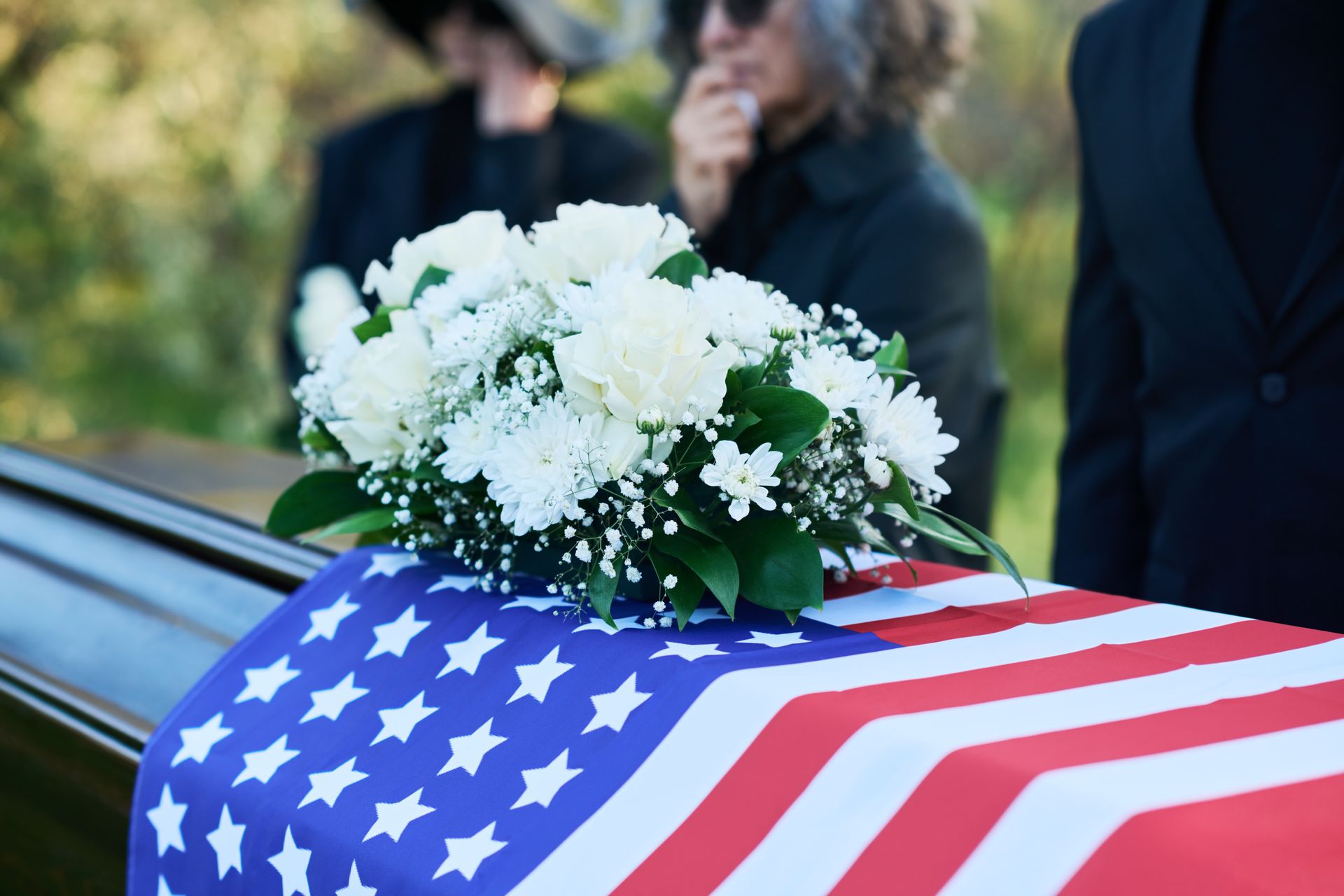Coffin draped in American flag with white flowers; mourners in background.