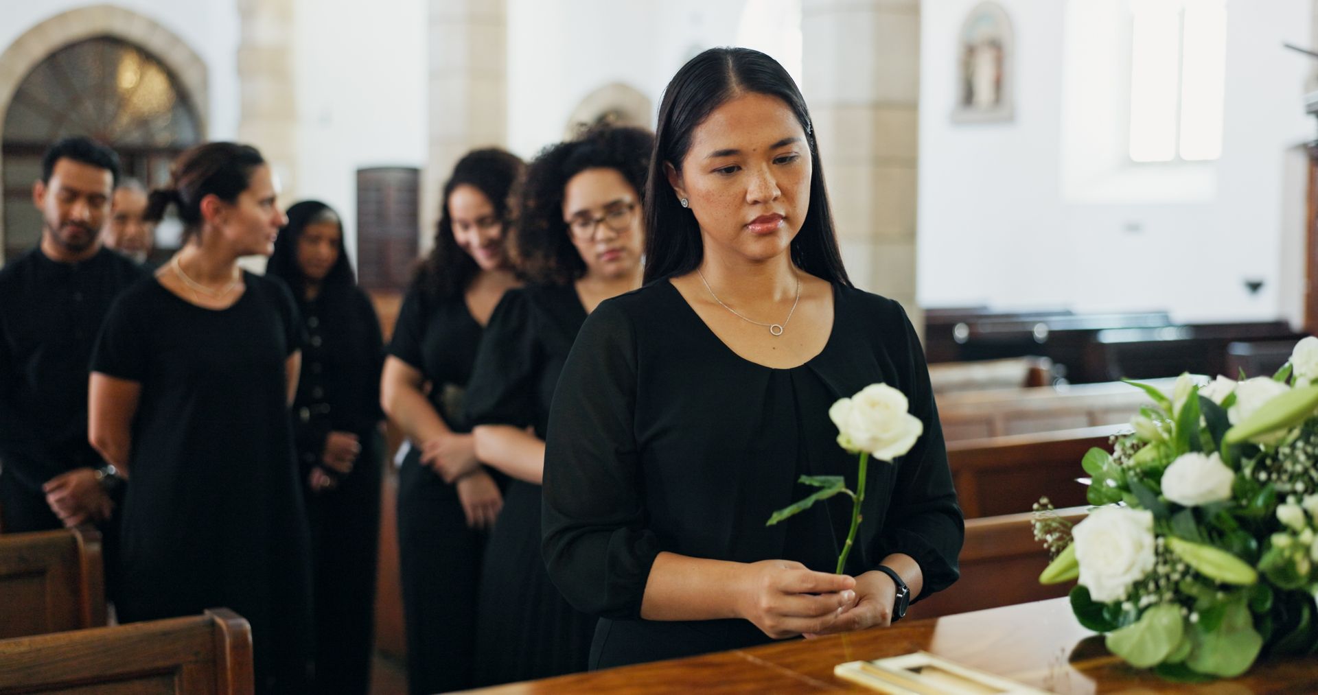 People in black at a funeral, woman holding a white rose, standing by a casket in a church.