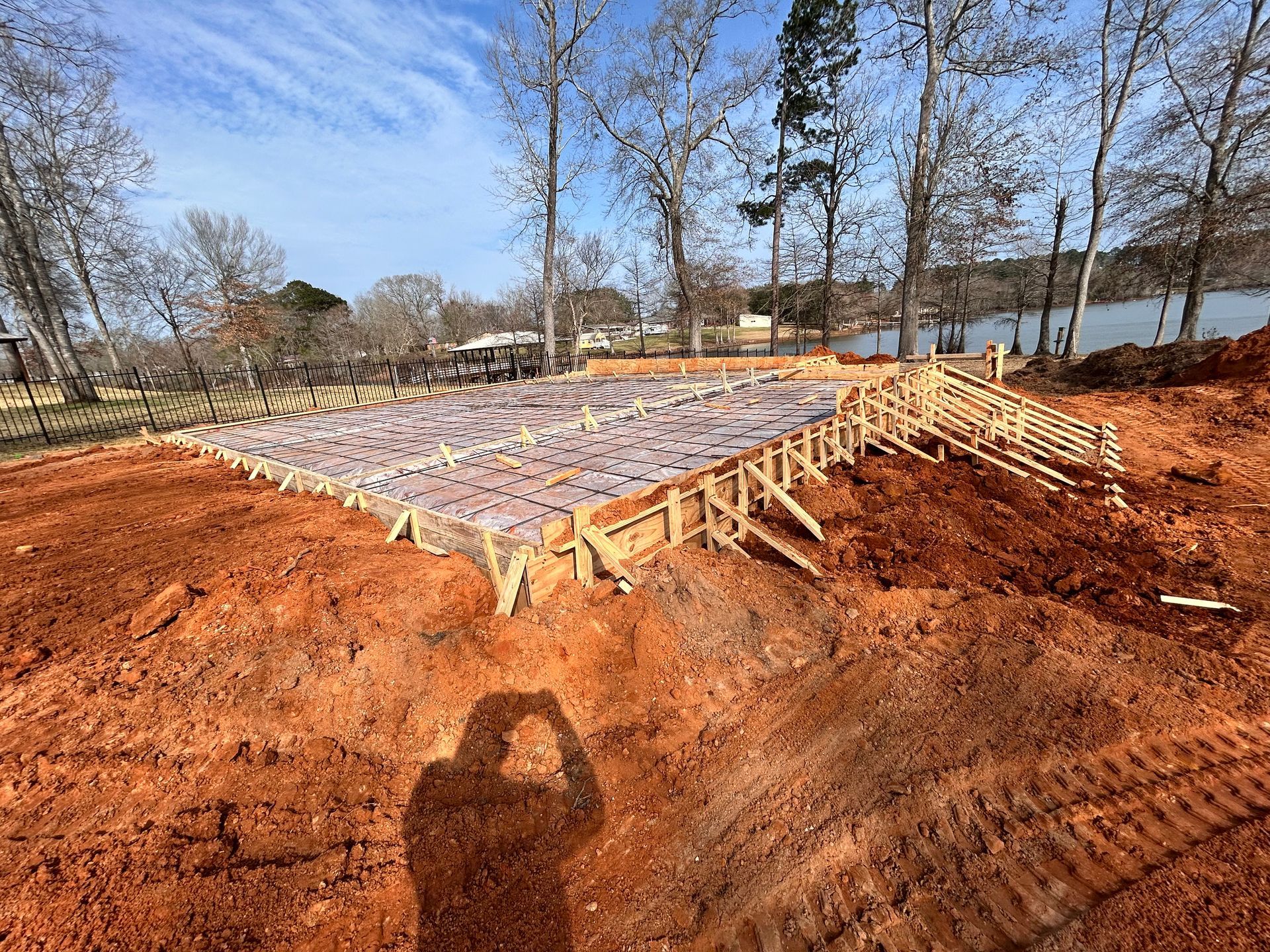 A wooden form for a concrete slab foundation sits in a cleared lot with red clay soil, under a clear blue sky.