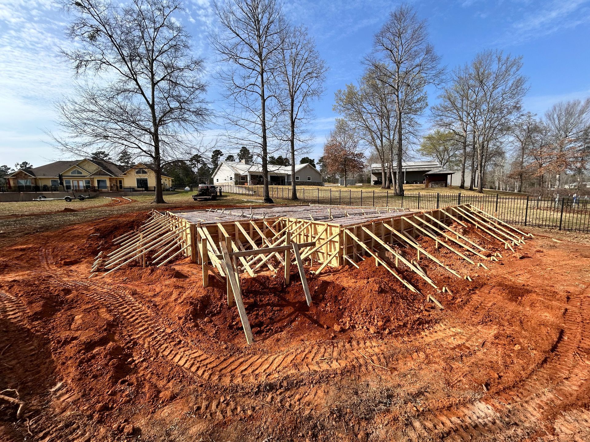 Wooden framework for a concrete foundation sits in a dirt-filled lot surrounded by trees and houses under a blue sky.