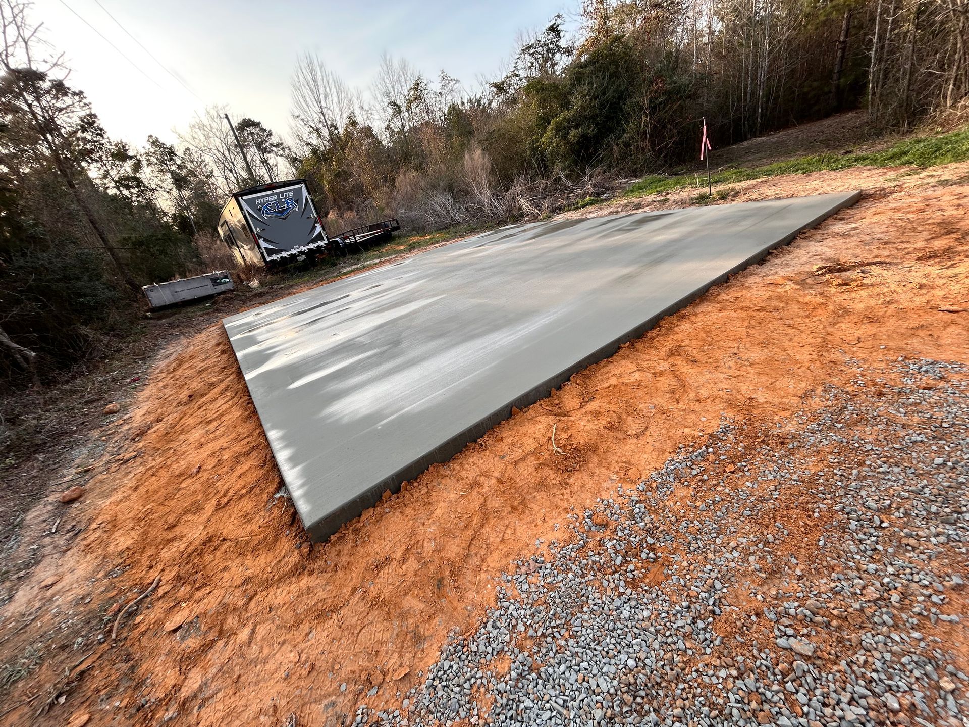 A freshly poured rectangular concrete slab surrounded by dirt and gravel in a wooded area.