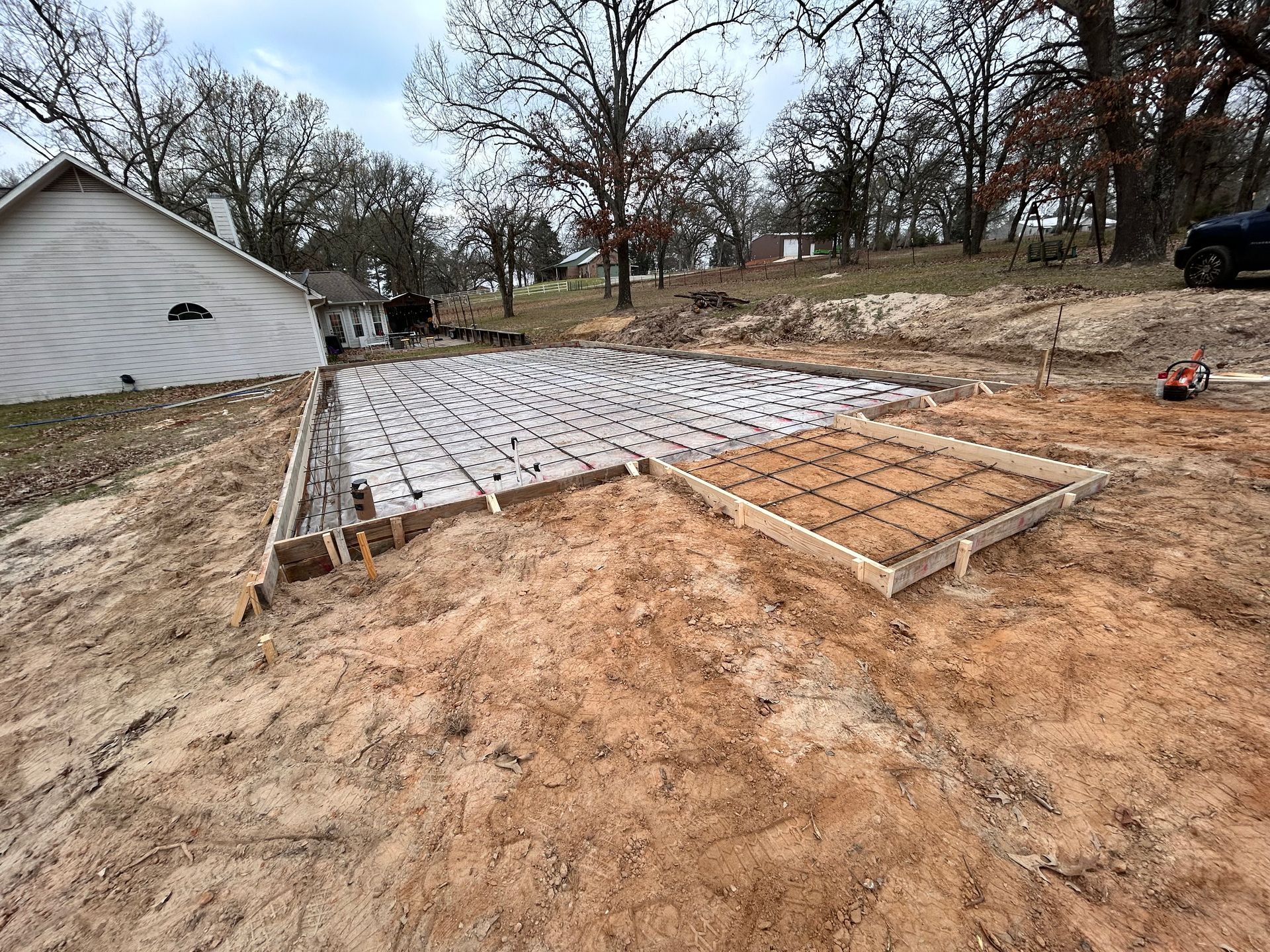 A construction site showing a concrete slab foundation prepared with rebar grids and plumbing pipes next to a white house.