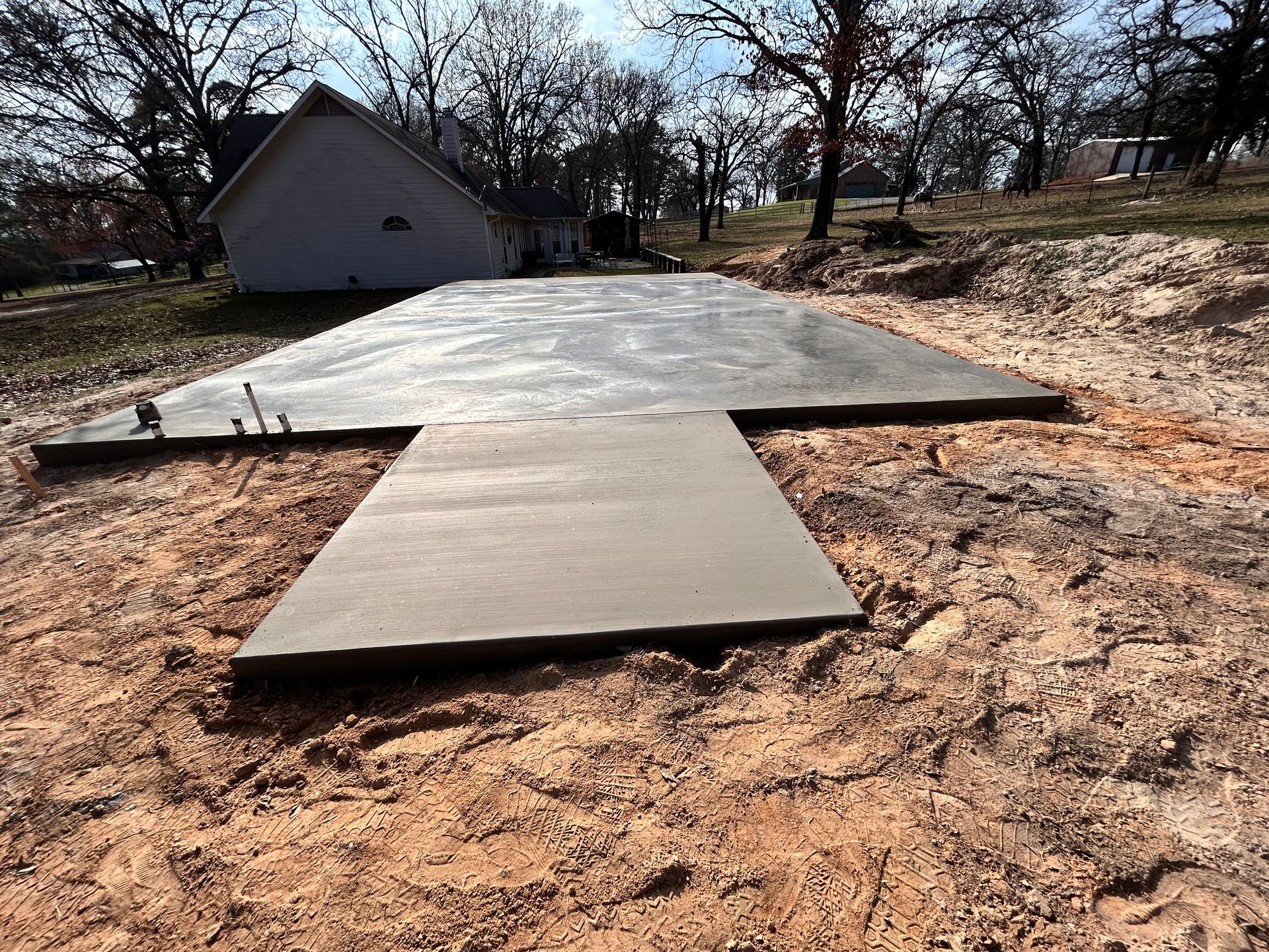 A newly poured concrete slab foundation sits in a dirt clearing with a small house in the background.