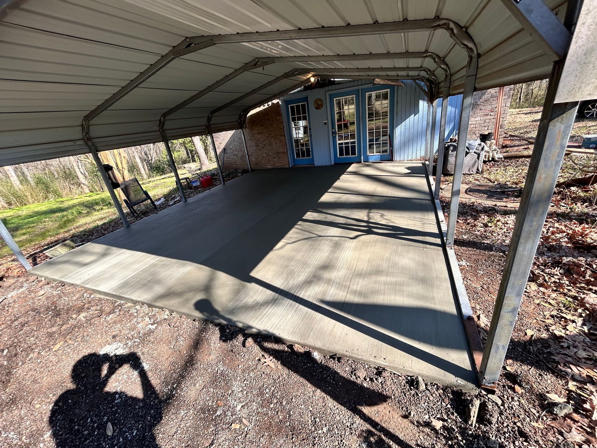 Carport with concrete slab, blue building, and trees. Shadow of person taking photo.