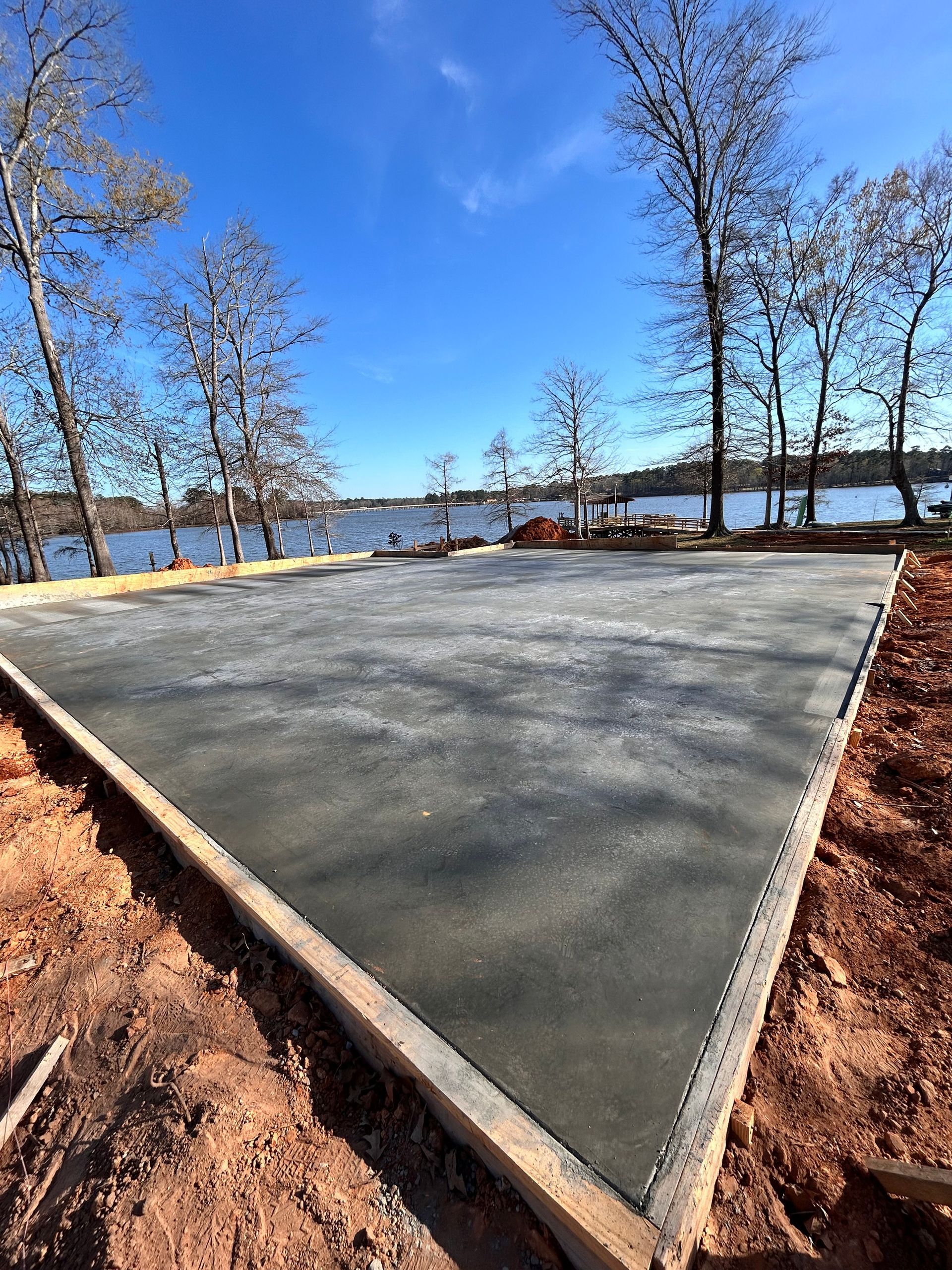 A freshly poured concrete foundation slab sits on a dirt lot overlooking a lake under a clear blue sky.