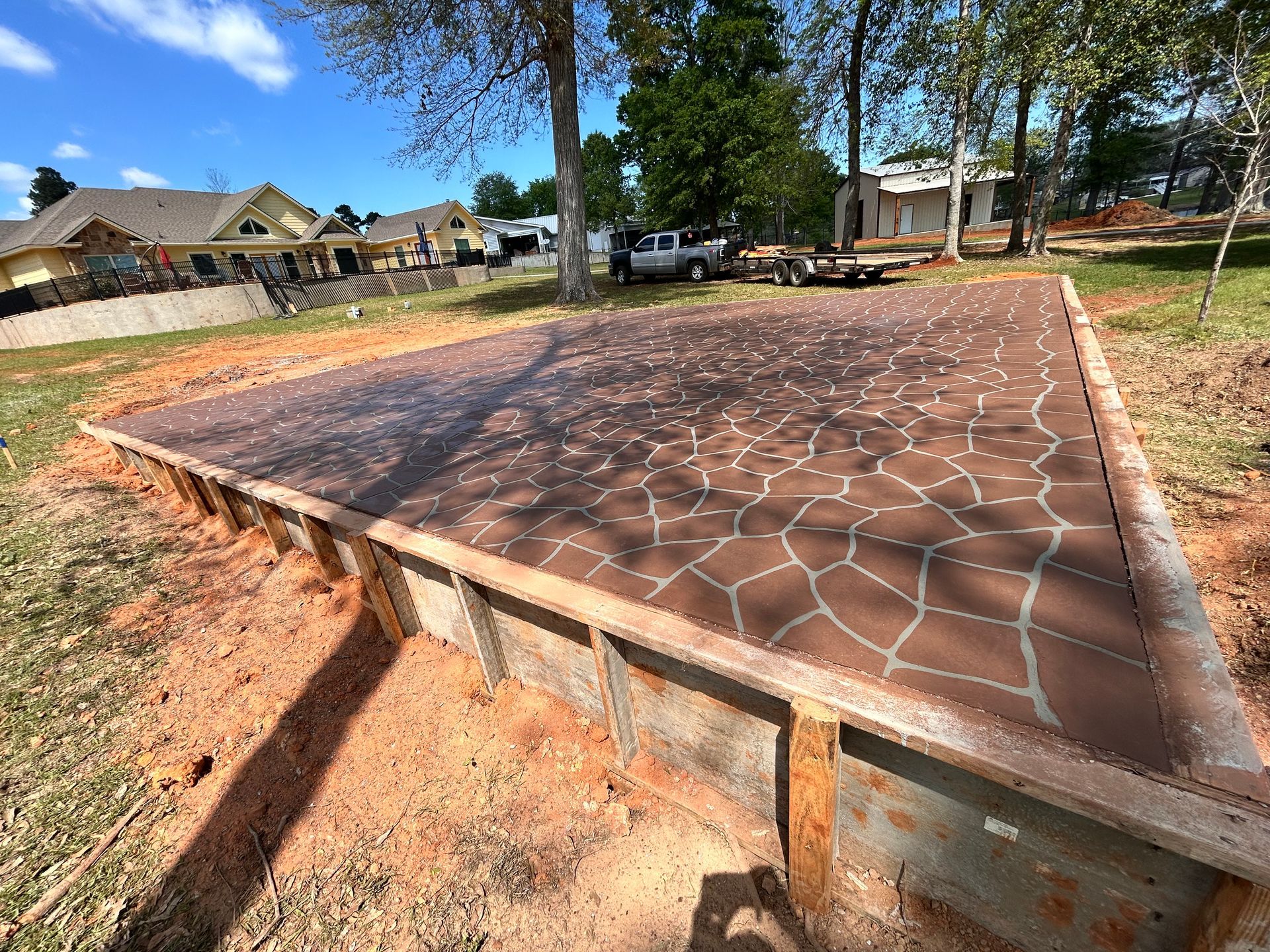 A newly poured concrete patio slab with a decorative flagstone pattern, surrounded by wooden forms on a dirt lot.