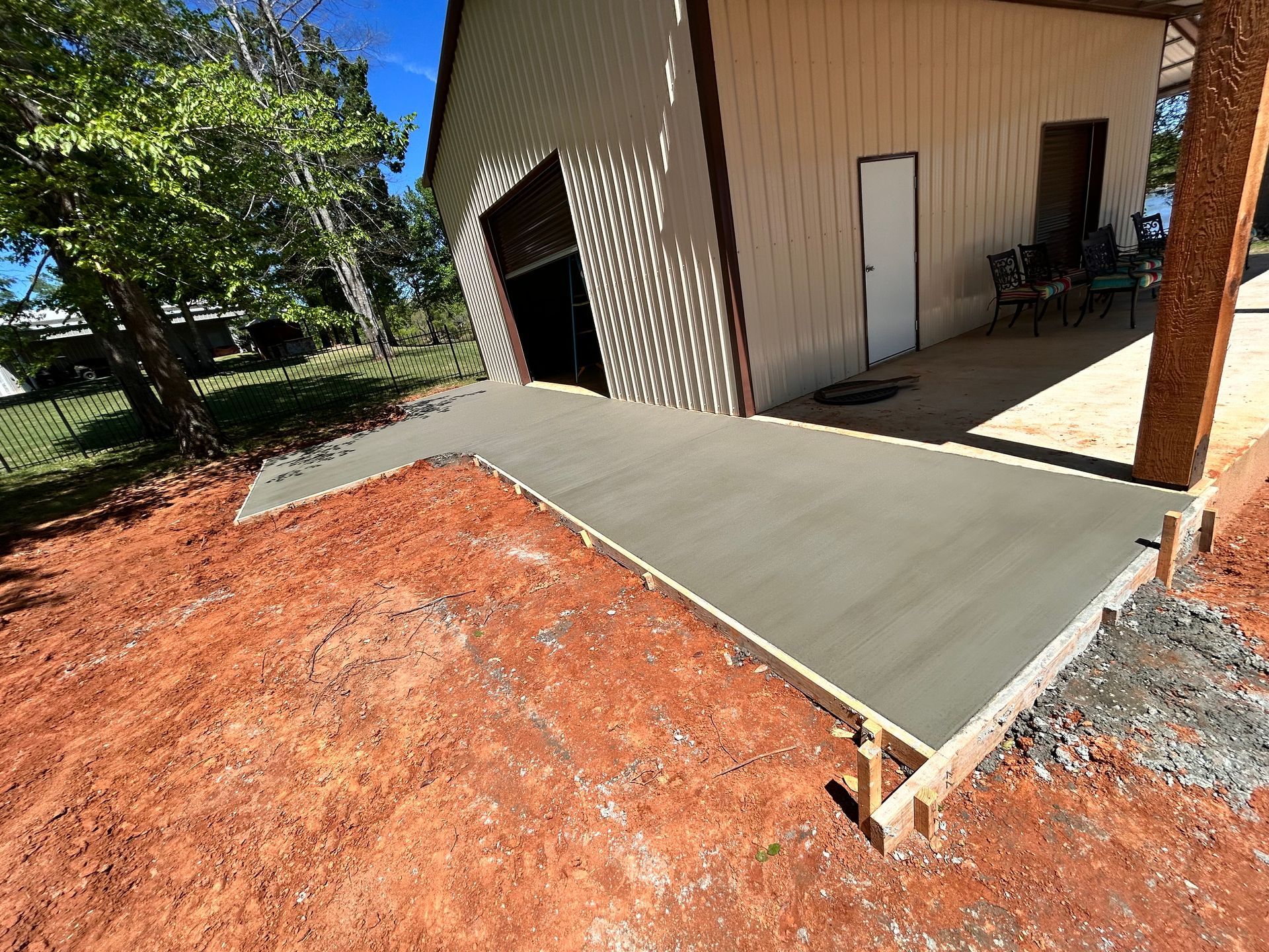 A fresh, smooth concrete sidewalk poured alongside a tan metal building on a dirt lot under a clear blue sky.