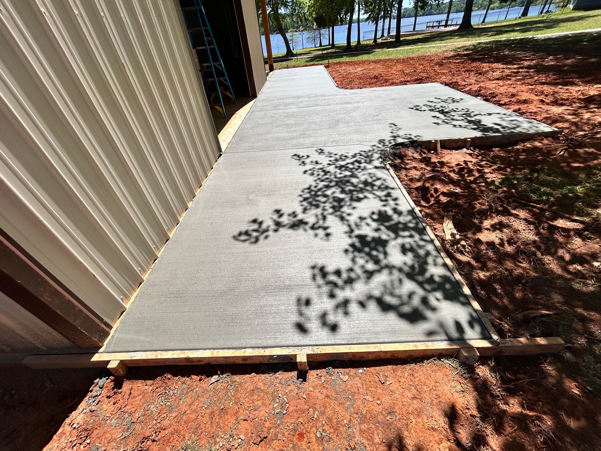 Newly poured concrete slab adjacent to a metal building, with wooden formwork and red soil nearby.