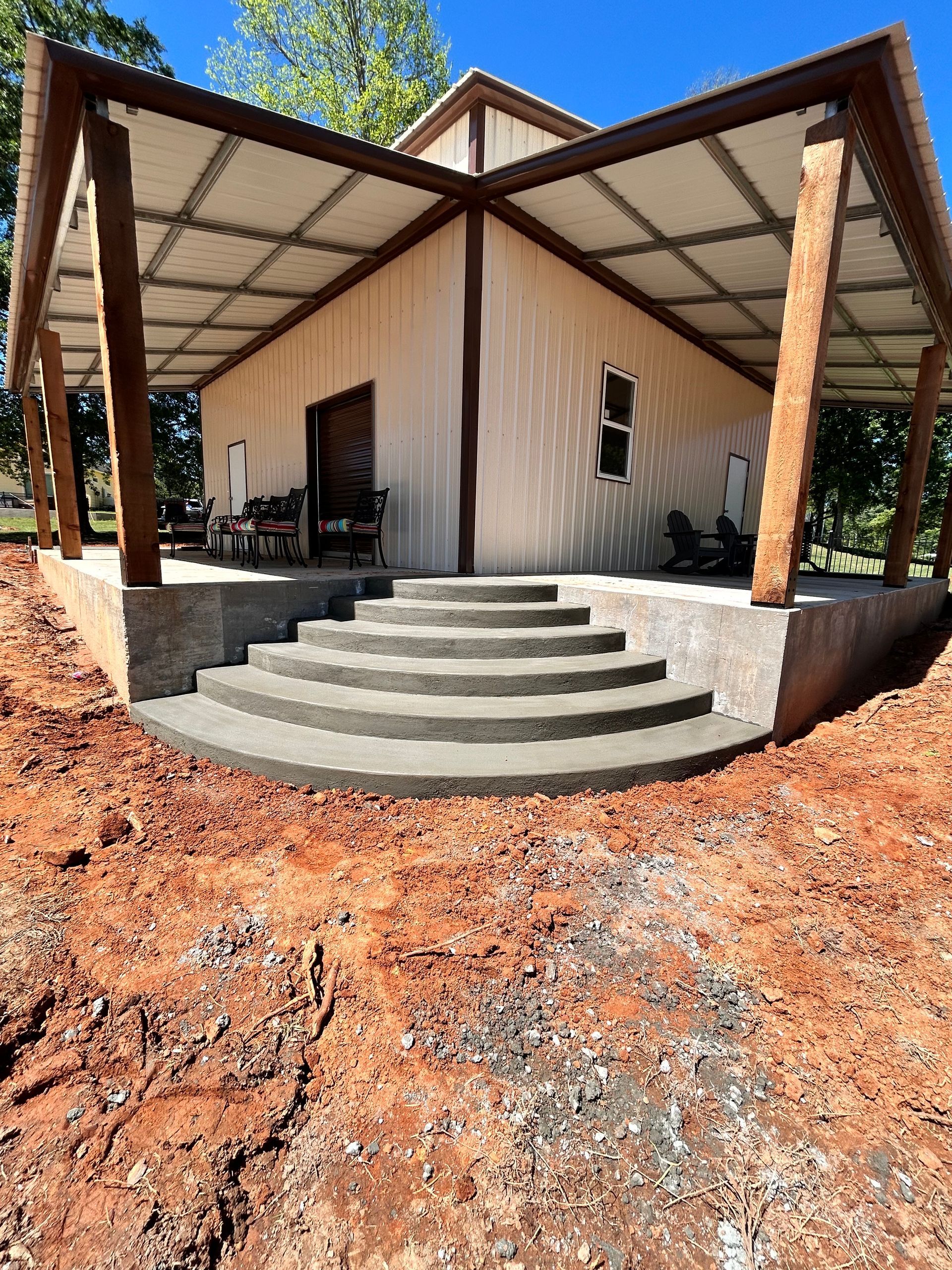 A small building with a tan metal exterior, wraparound porch, and semi-circular concrete stairs on a dirt construction site.