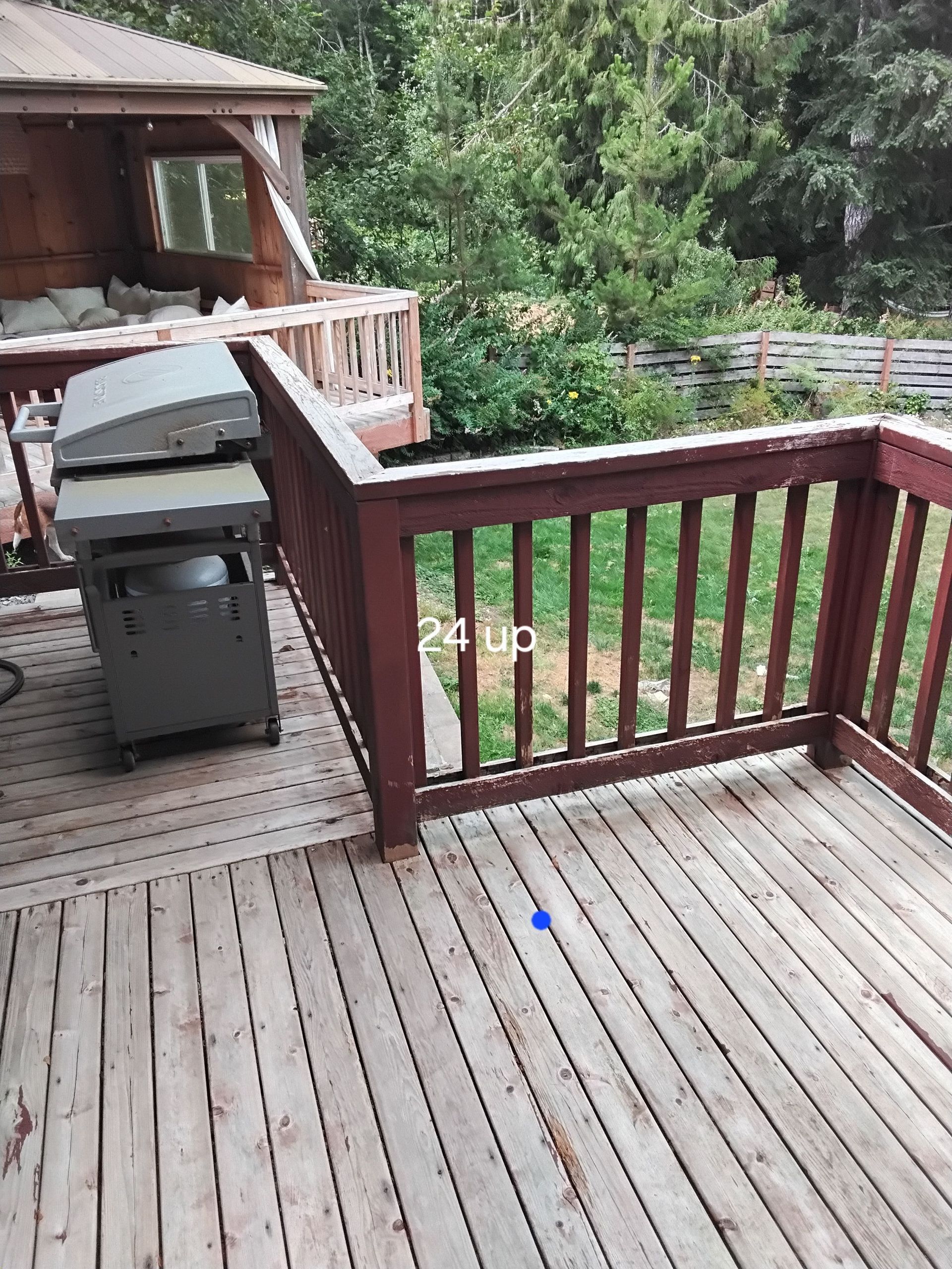Wooden deck with grill, railing, and a cabin in the background, surrounded by trees.