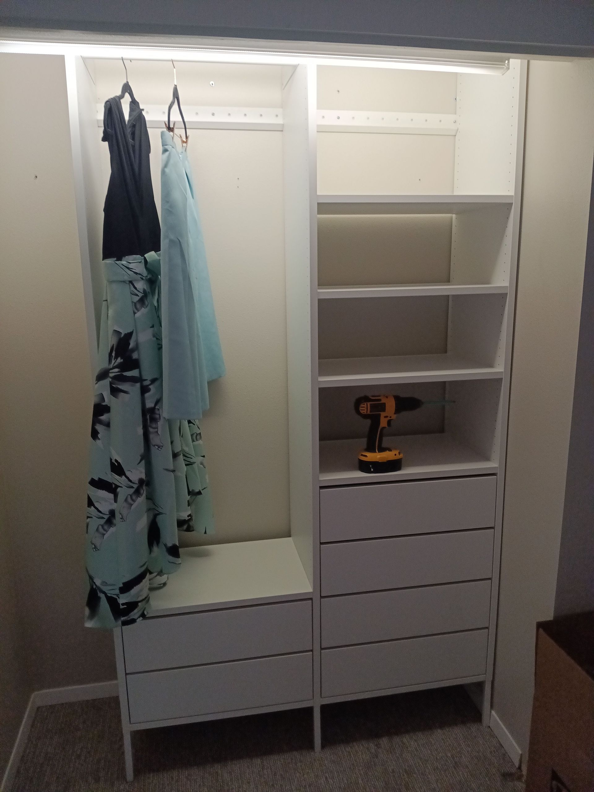Built-in white closet with hanging clothes, shelves, and drawers. A power drill rests on a shelf.