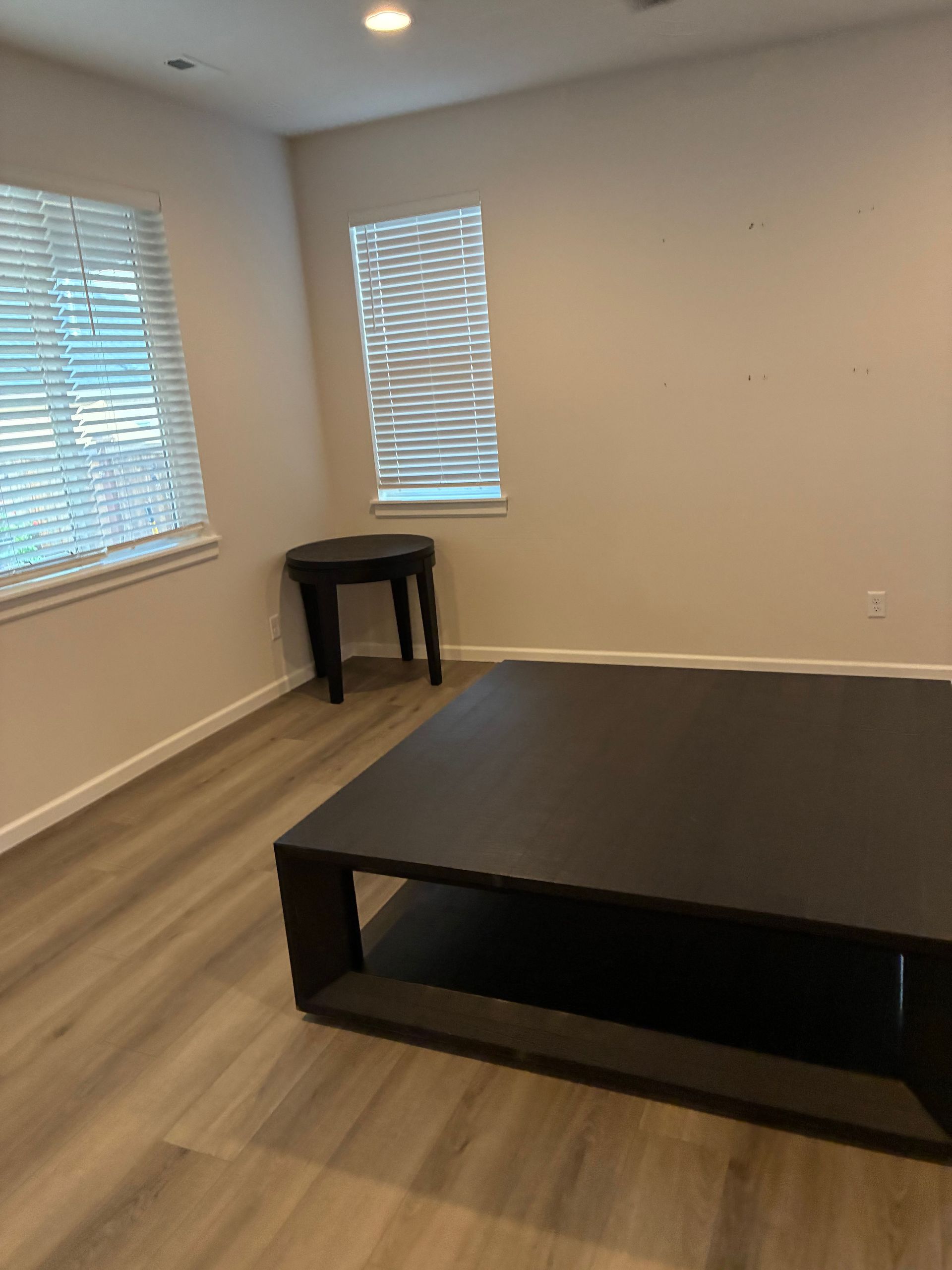 Empty room with dark brown coffee table, small side table, two windows with blinds, and light colored flooring.