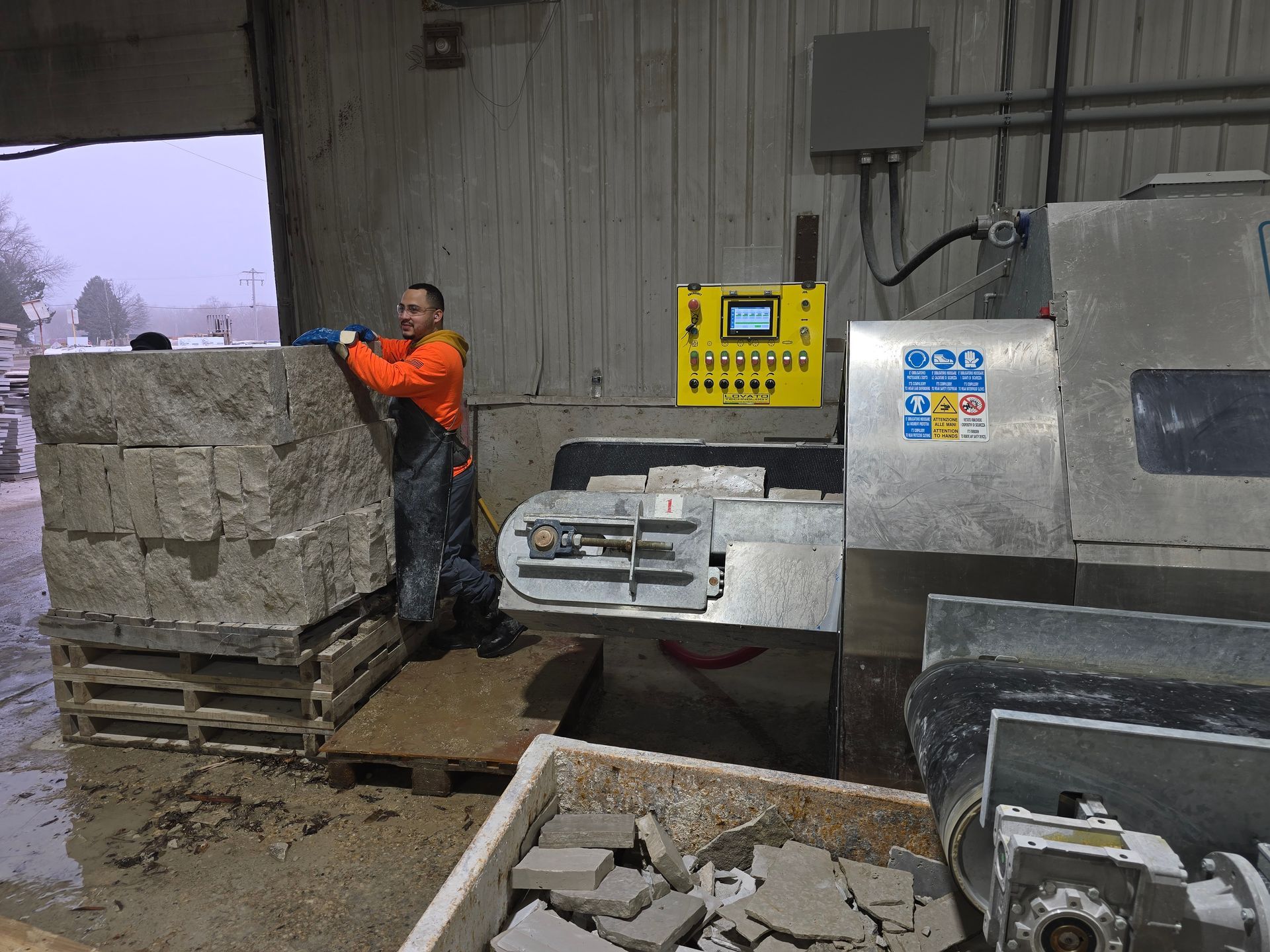 A person in work gear near a stone-cutting machine inside a gray industrial building.
