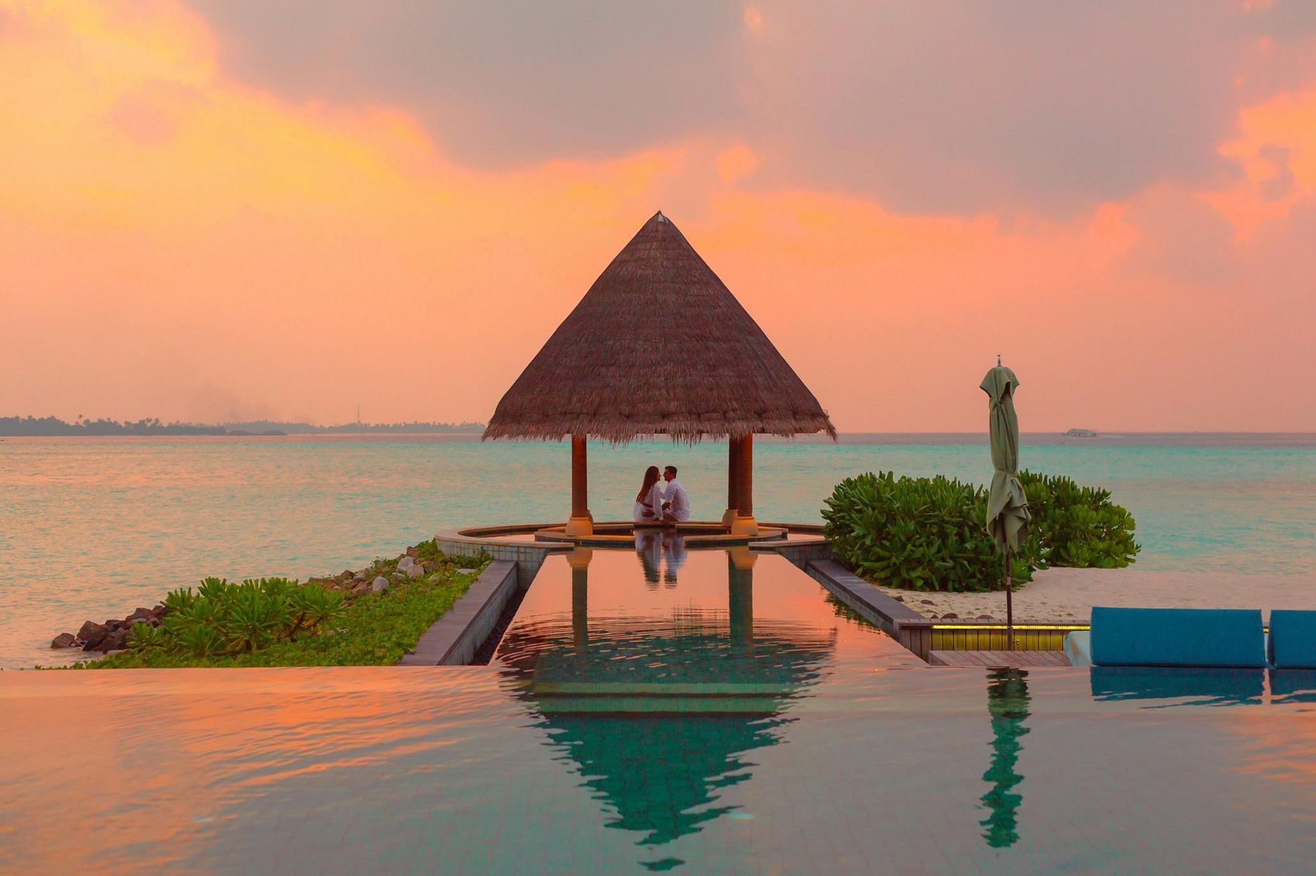 A couple is sitting under a thatched hut next to an infinity pool.