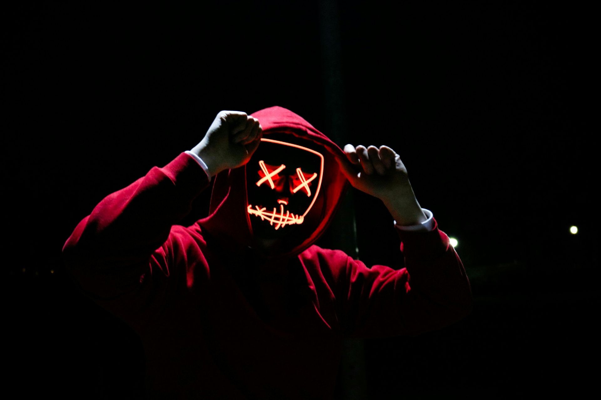 A person wearing a red hoodie with a neon mask on their face.