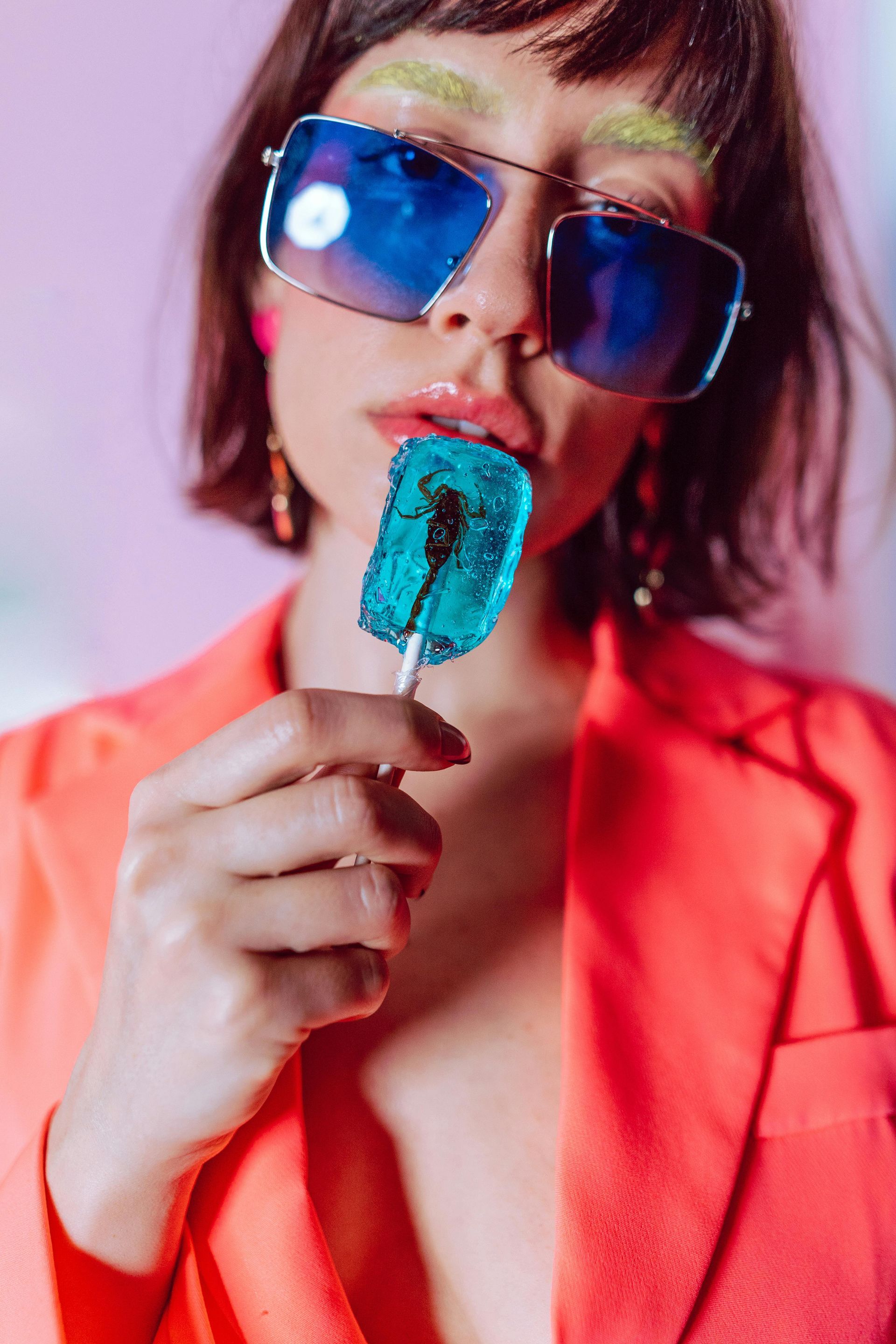 A woman wearing sunglasses is holding a blue lollipop in her hand.