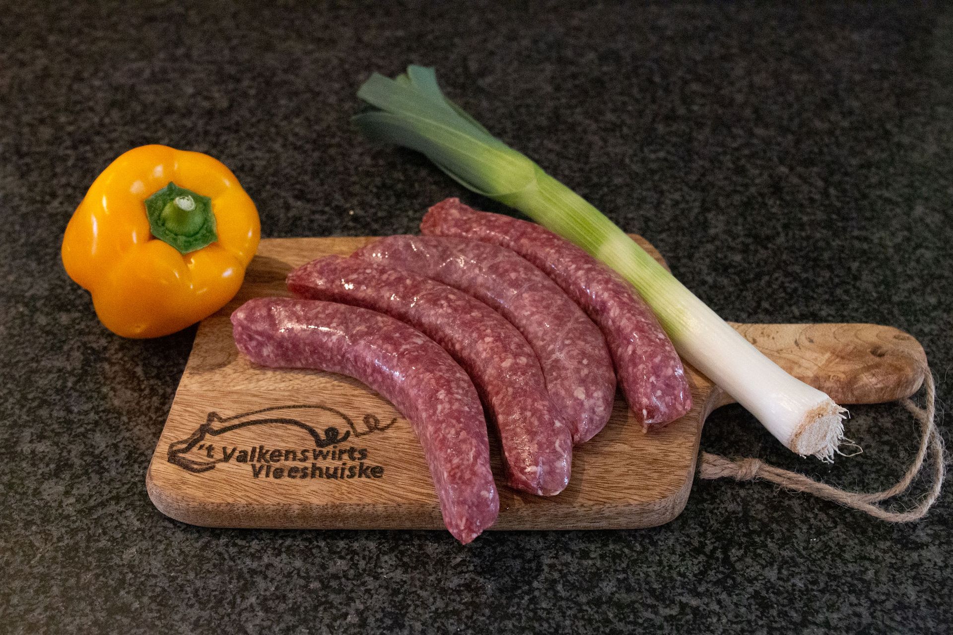 Gele paprika, rauwe worstjes en prei op een houten snijplank.