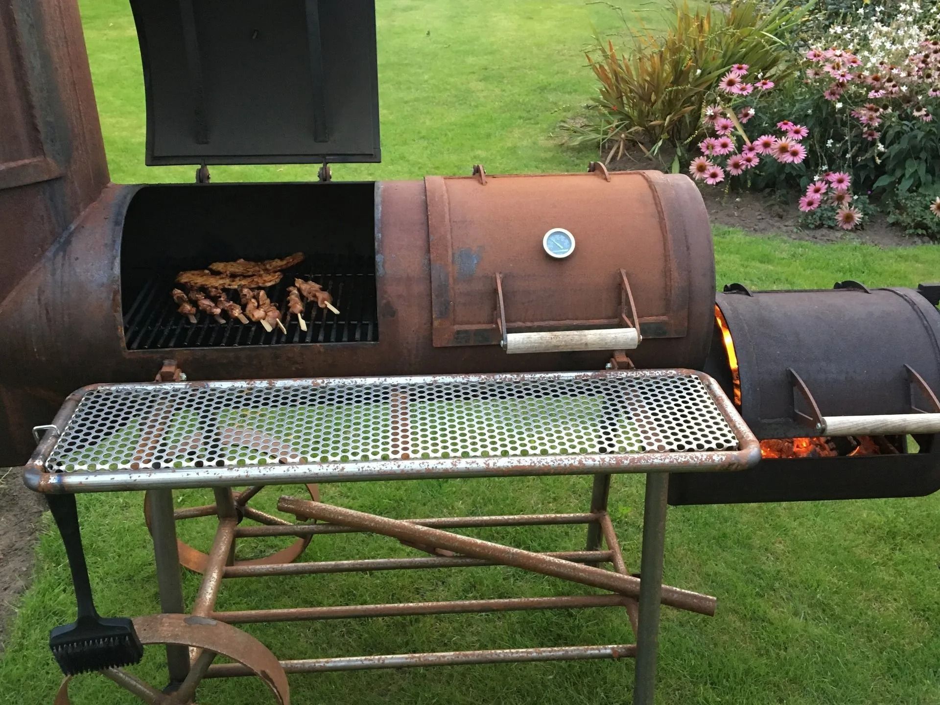 Roestige metalen roker die spiesjes grilt op een groen gazon met bloemen.
