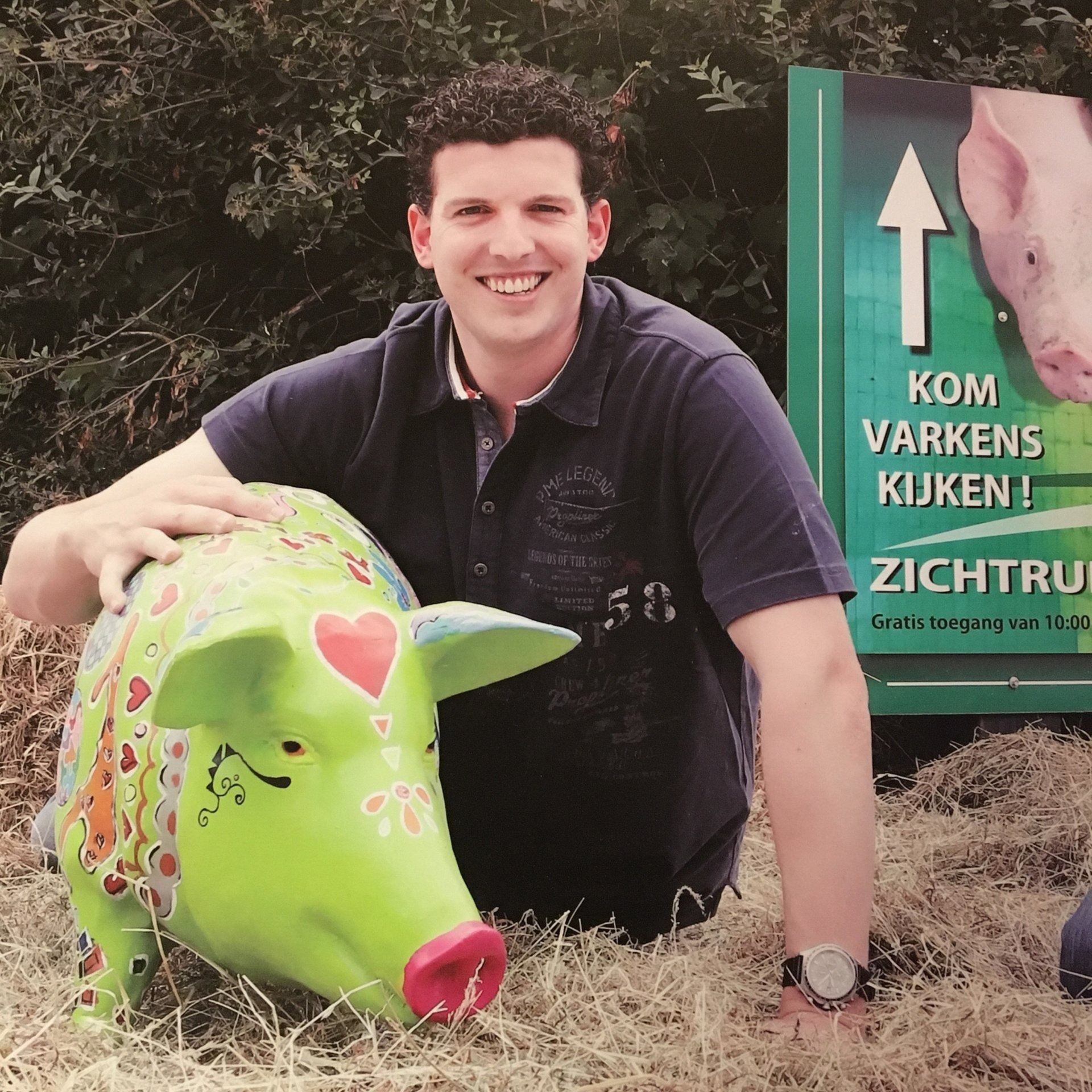 Man lacht en hurkt naast een groen varkensbeeld met een hartmotief. Het bordje met de pijl wijst naar boven.