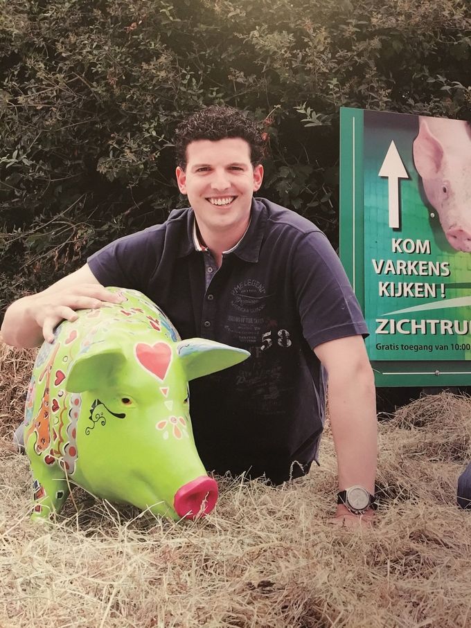 Man lacht en hurkt naast een groen varkensbeeld met een hartmotief. Het bordje met de pijl wijst naar boven.